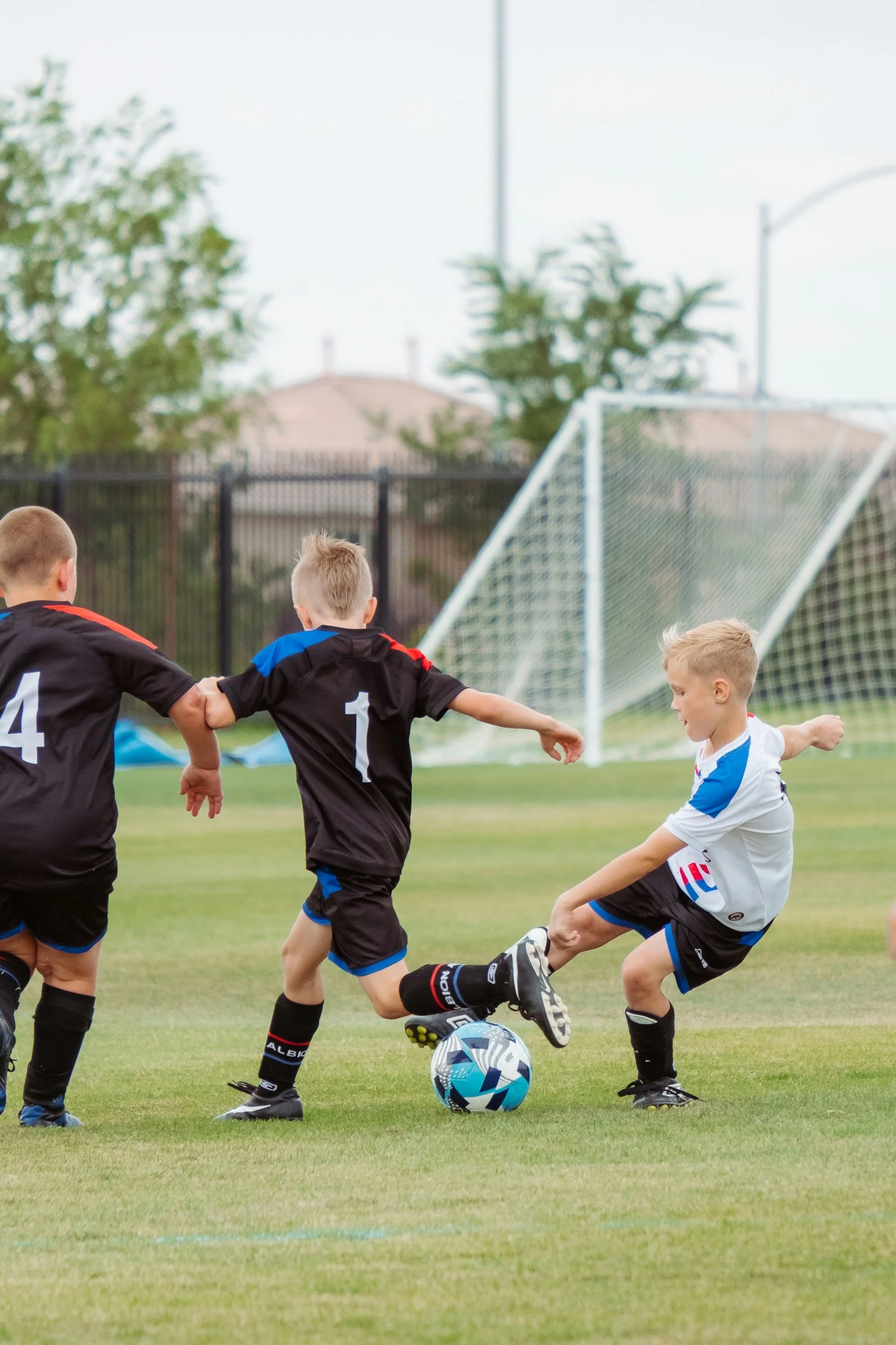 Young boys playing soccer on a field, with one kicking the ball while others attempt to block or intercept.