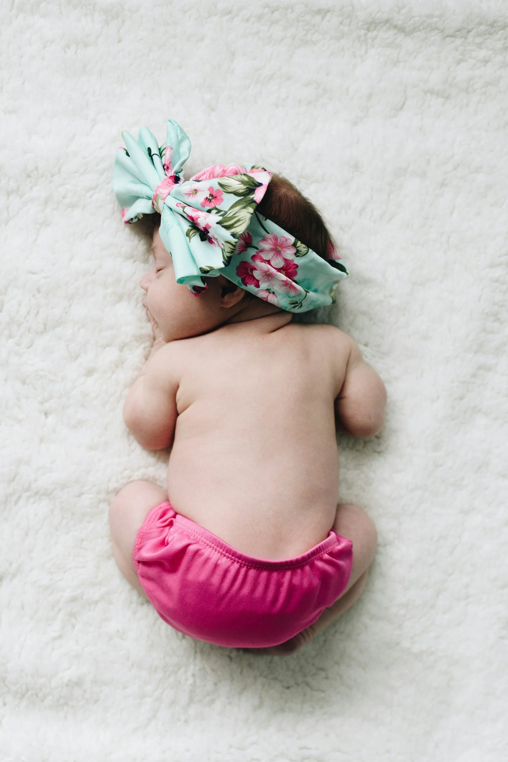A sleeping baby lying face down on a soft white fuzzy blanket, wearing a pink diaper and a large colorful floral headband.