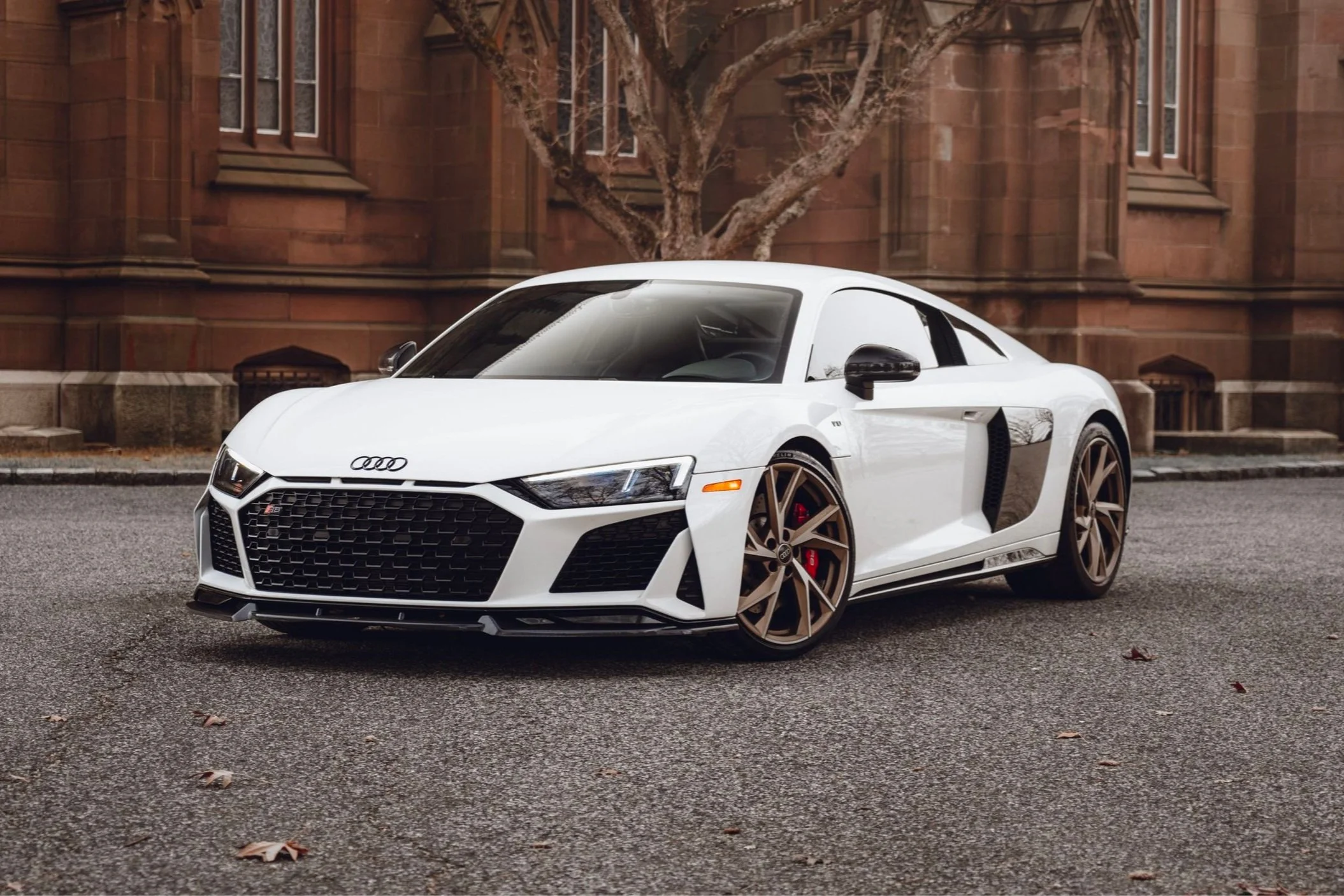 A white Audi R8 sports car parked on a city street with brown brick buildings and a tree in the background.