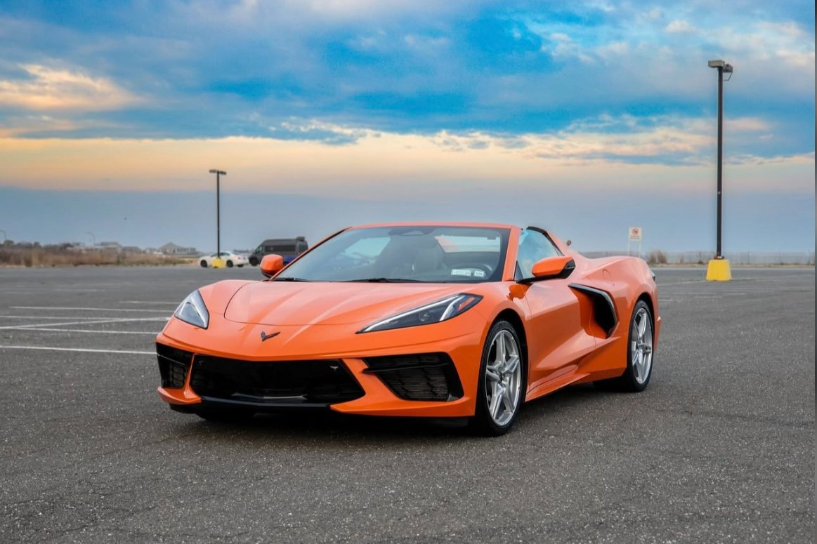 An orange Chevrolet Corvette sports car parked in an empty parking lot under a partly cloudy sky.
