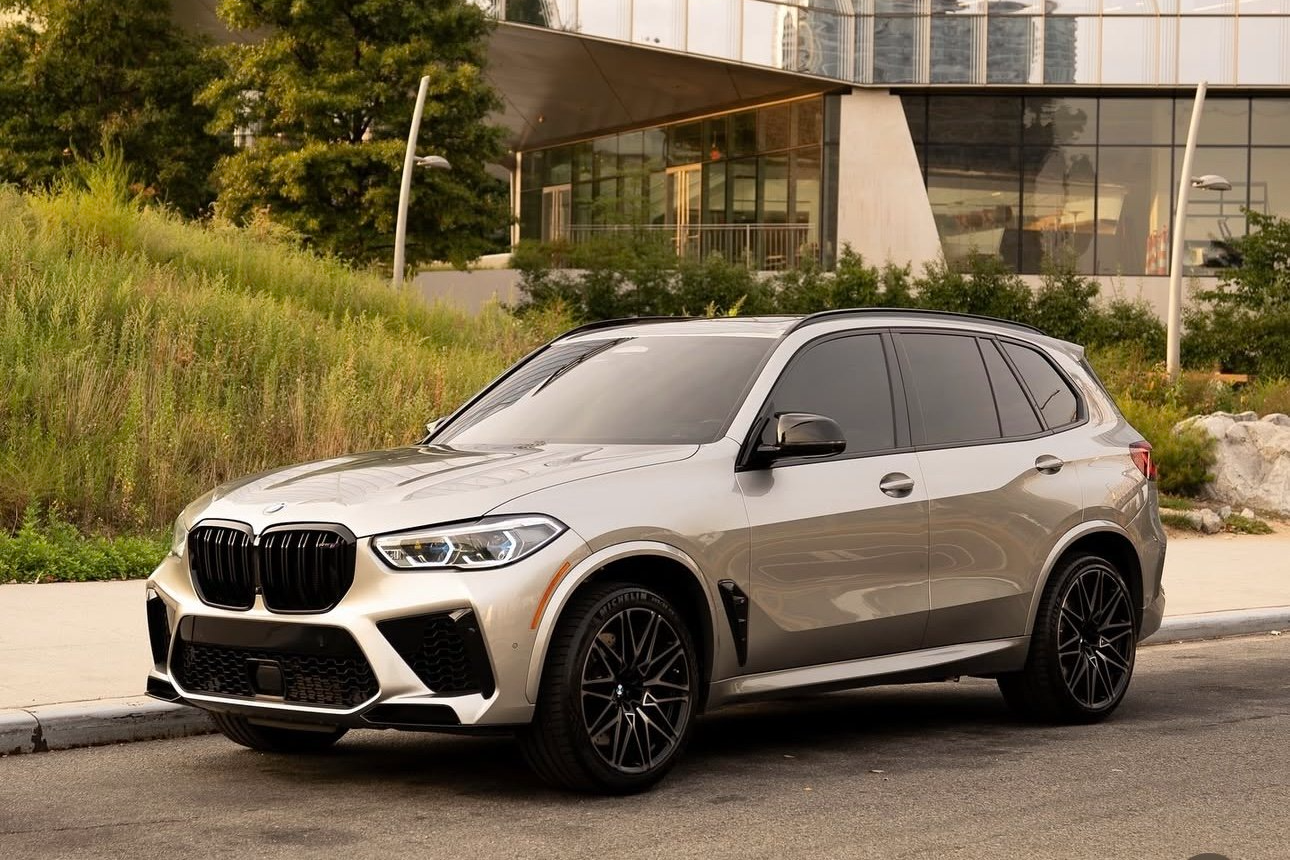 A silver BMW SUV parked on a city street with modern glass buildings and greenery nearby.