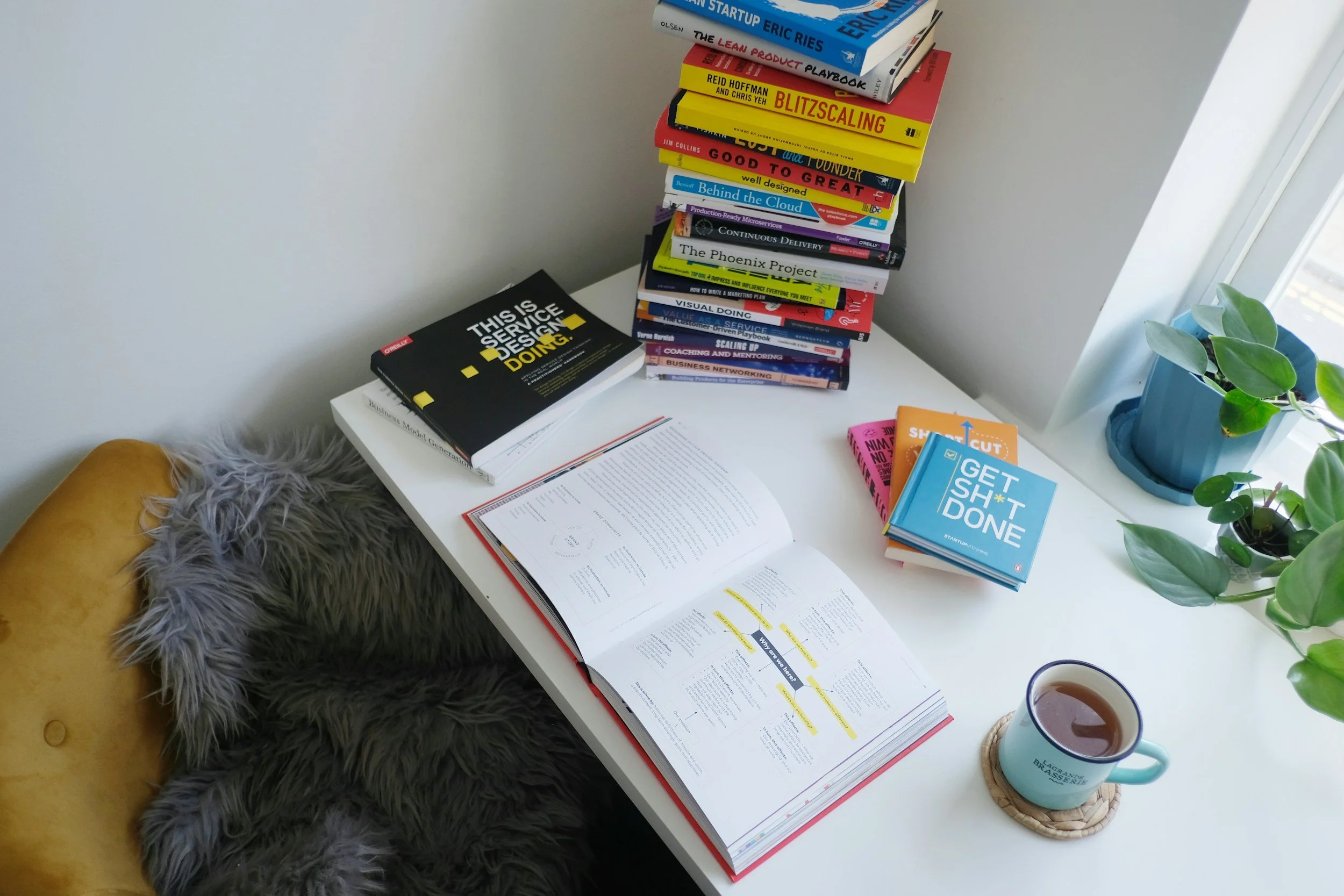 A white desk holds open study books, a stack of colorful books, a blue mug filled with coffee on a coaster, and a small potted plant with green leaves near a window. A chair with a yellow cushion and gray fur throw is beside the desk.