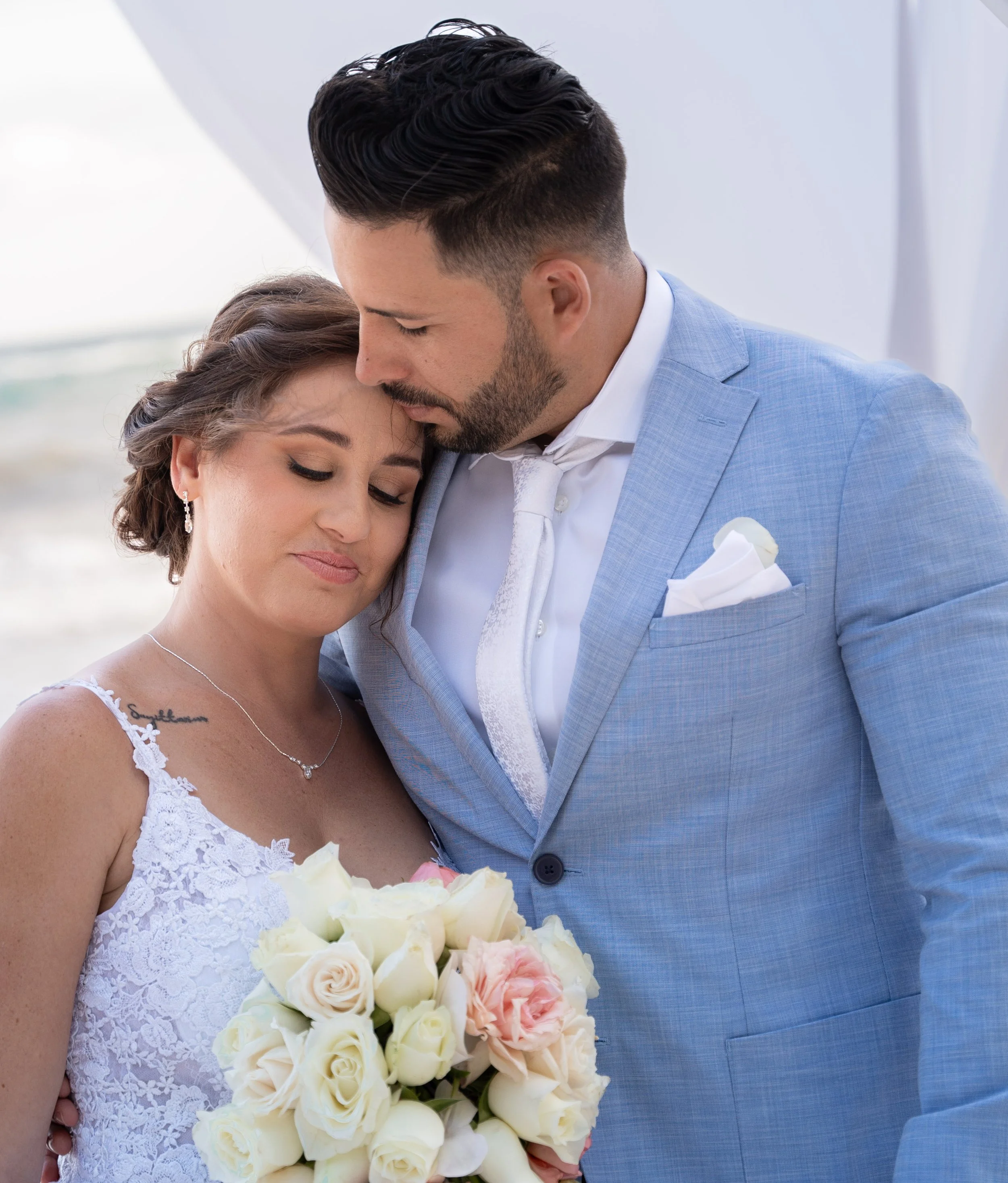A bride and groom share an intimate moment on the beach, with the groom gently resting his forehead on the bride's head as she holds a bouquet of white and blush roses.