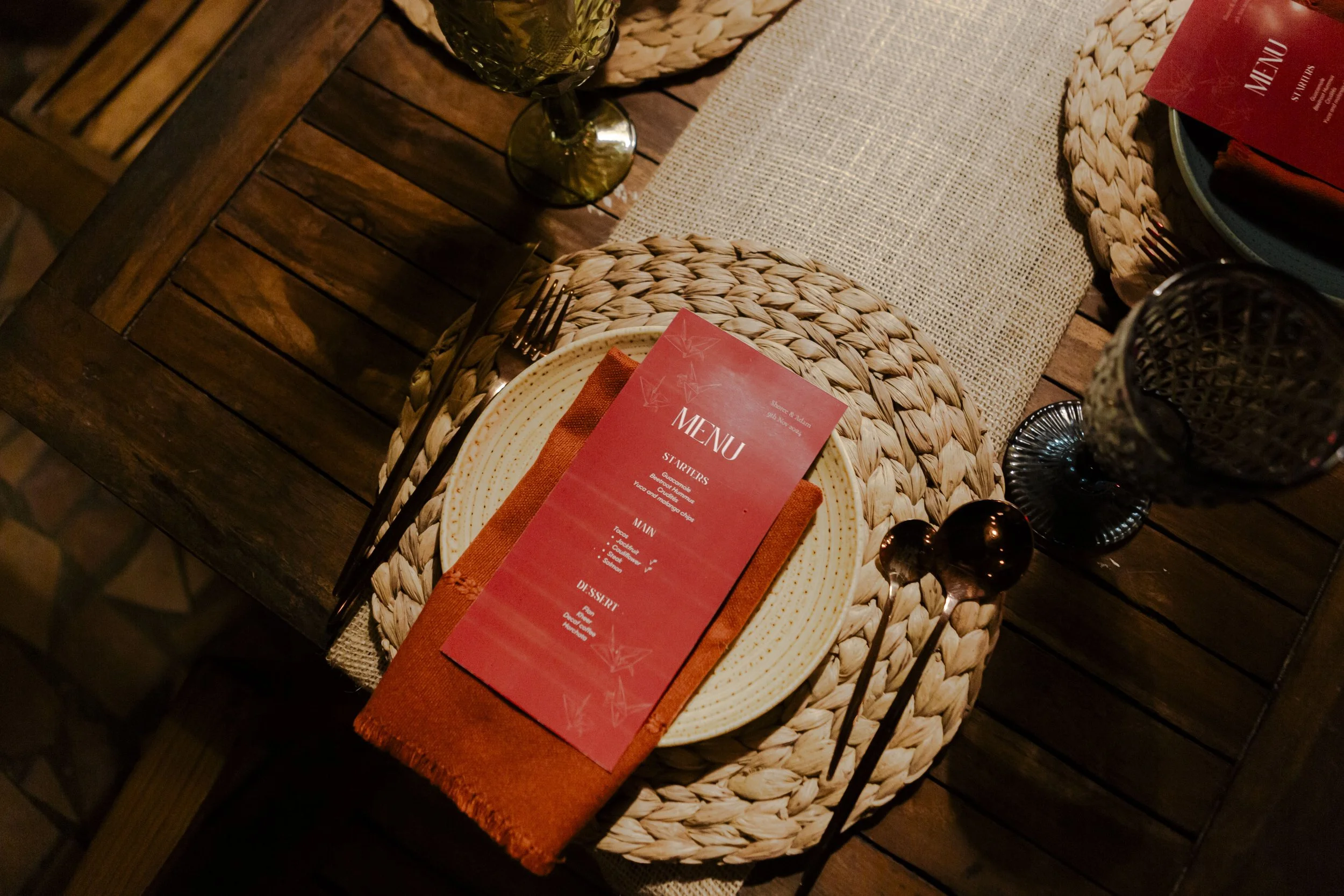 A table setting with a red menu on a cream-colored plate, woven placemat, glassware, and cutlery on a rustic wooden table.