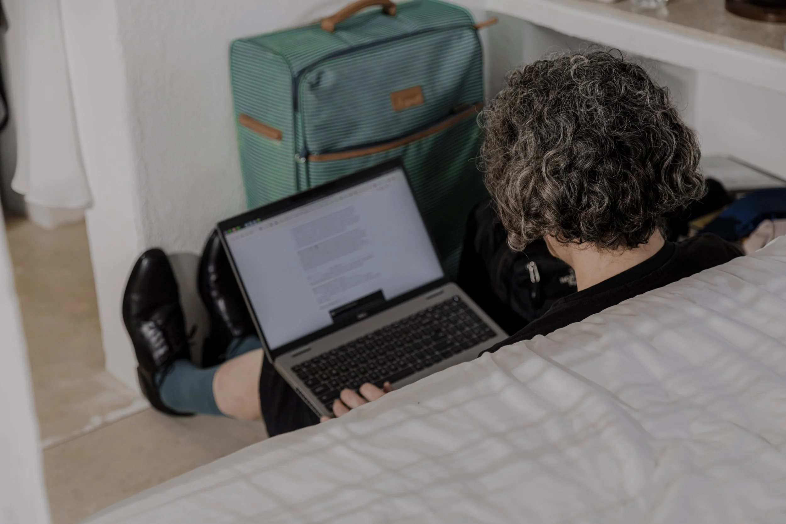 Person sitting on the floor with legs extended, working on a laptop, with a striped teal suitcase behind them and a bed in the foreground.