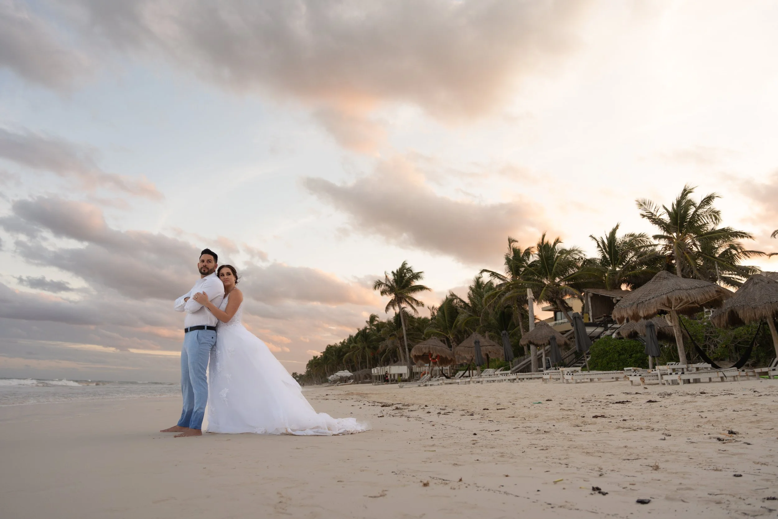 A bride in a white wedding dress and a groom in a white shirt and light blue pants standing arm-in-arm on a sandy beach at sunset with palm trees and beach umbrellas in the background.