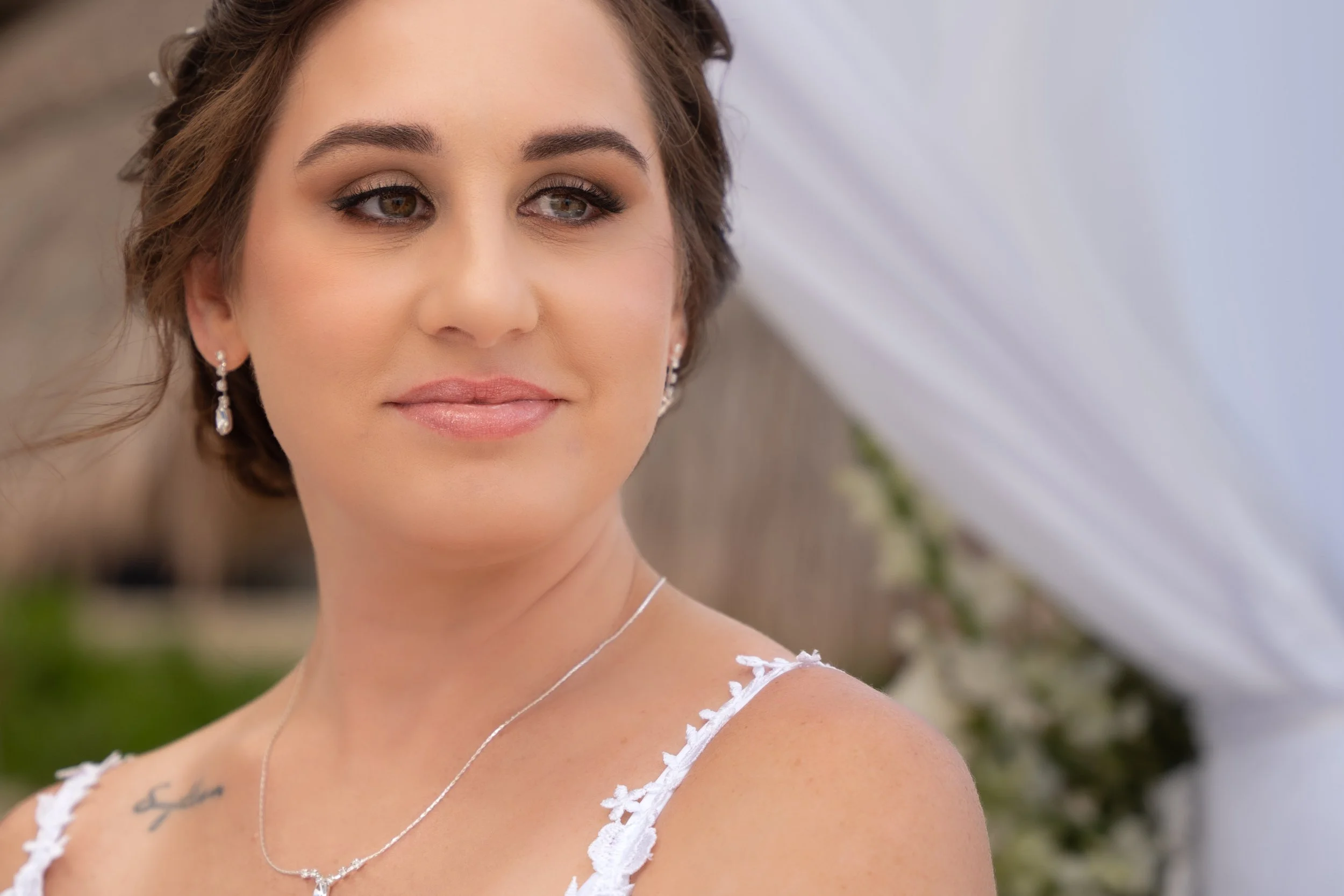 Close-up of a woman with brown hair and light makeup, wearing earrings and a delicate necklace, dressed in white with floral details, outdoors with a blurred background.