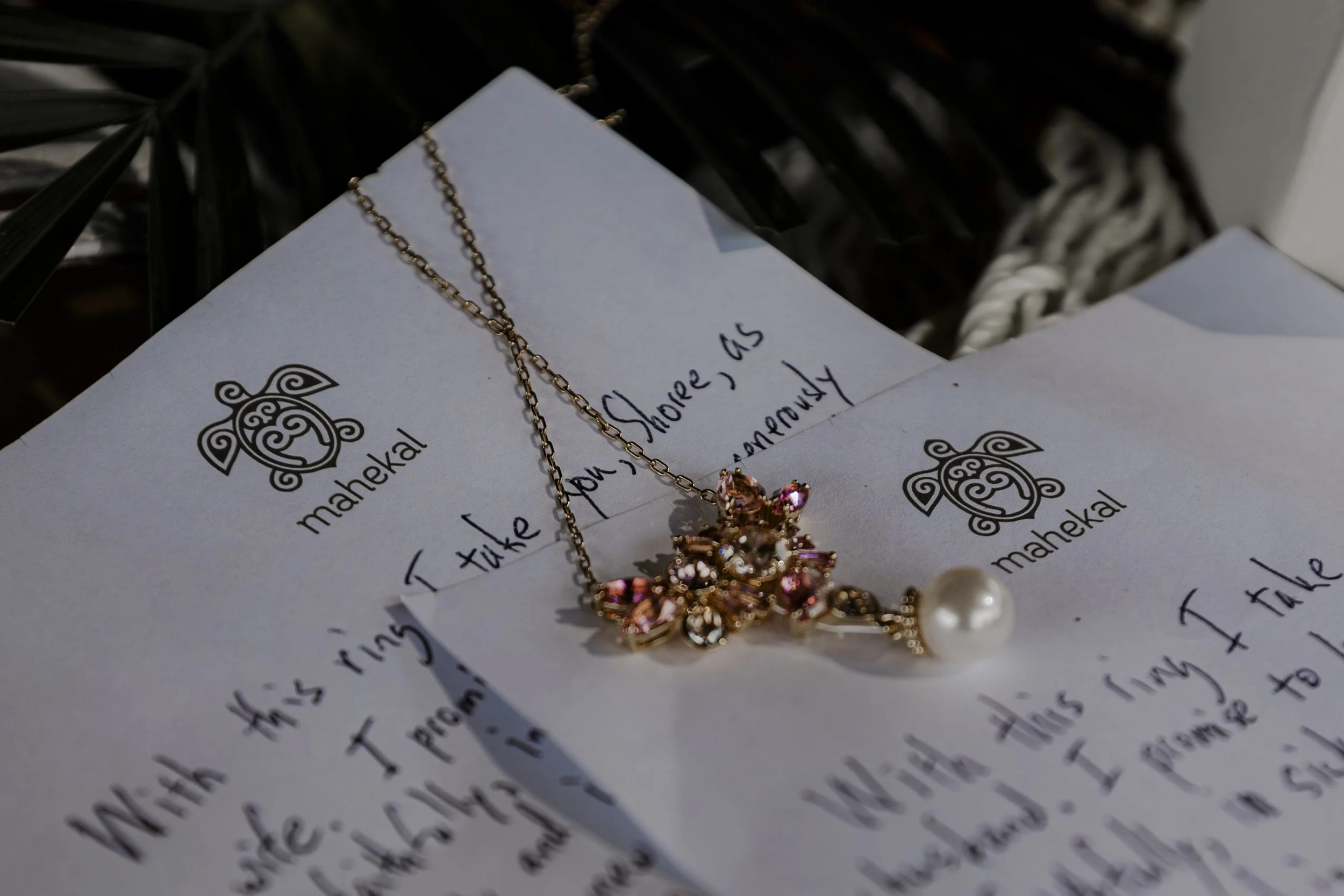 A gold necklace with pink and clear gemstones and a pearl charm resting on a handwritten letter on white paper with black text and a turtle logo.