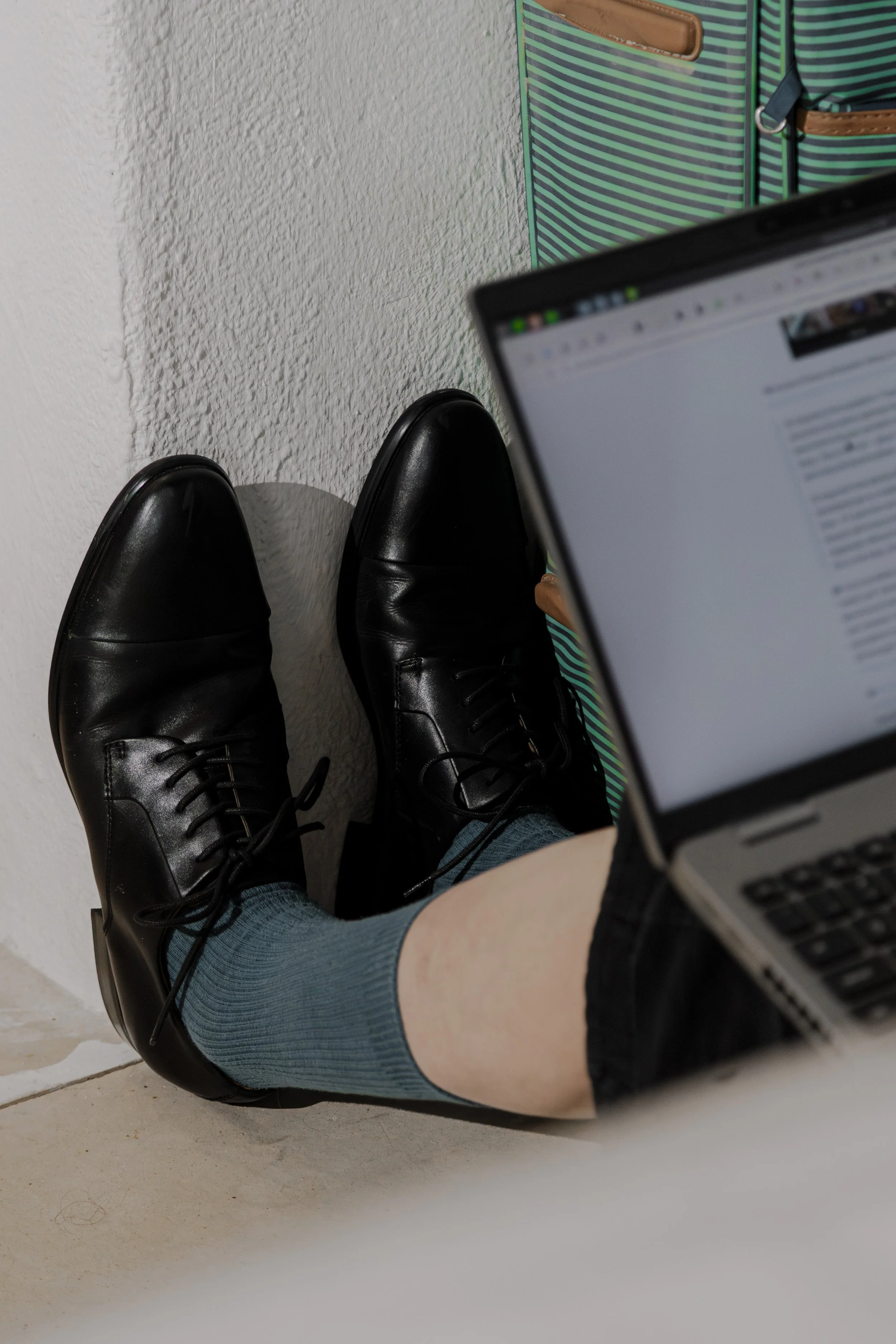 Person sitting on the floor with legs extended, wearing black dress shoes, blue socks, and black shorts, using a laptop. A striped bag hangs on the wall behind.