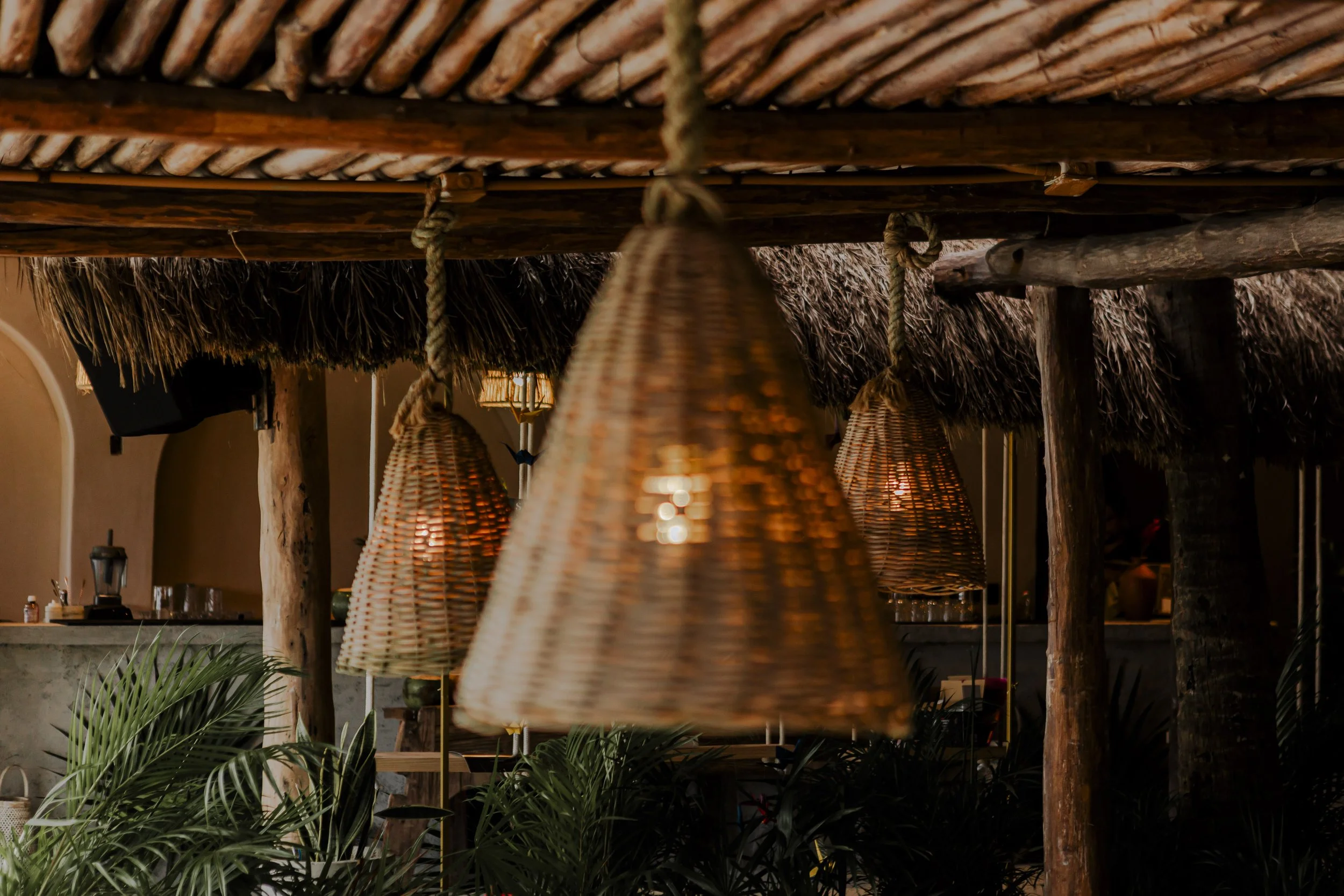Interior of a tropical-themed restaurant or bar with wicker pendant lights hanging from a thatched ceiling, wooden support beams, and tropical plants.