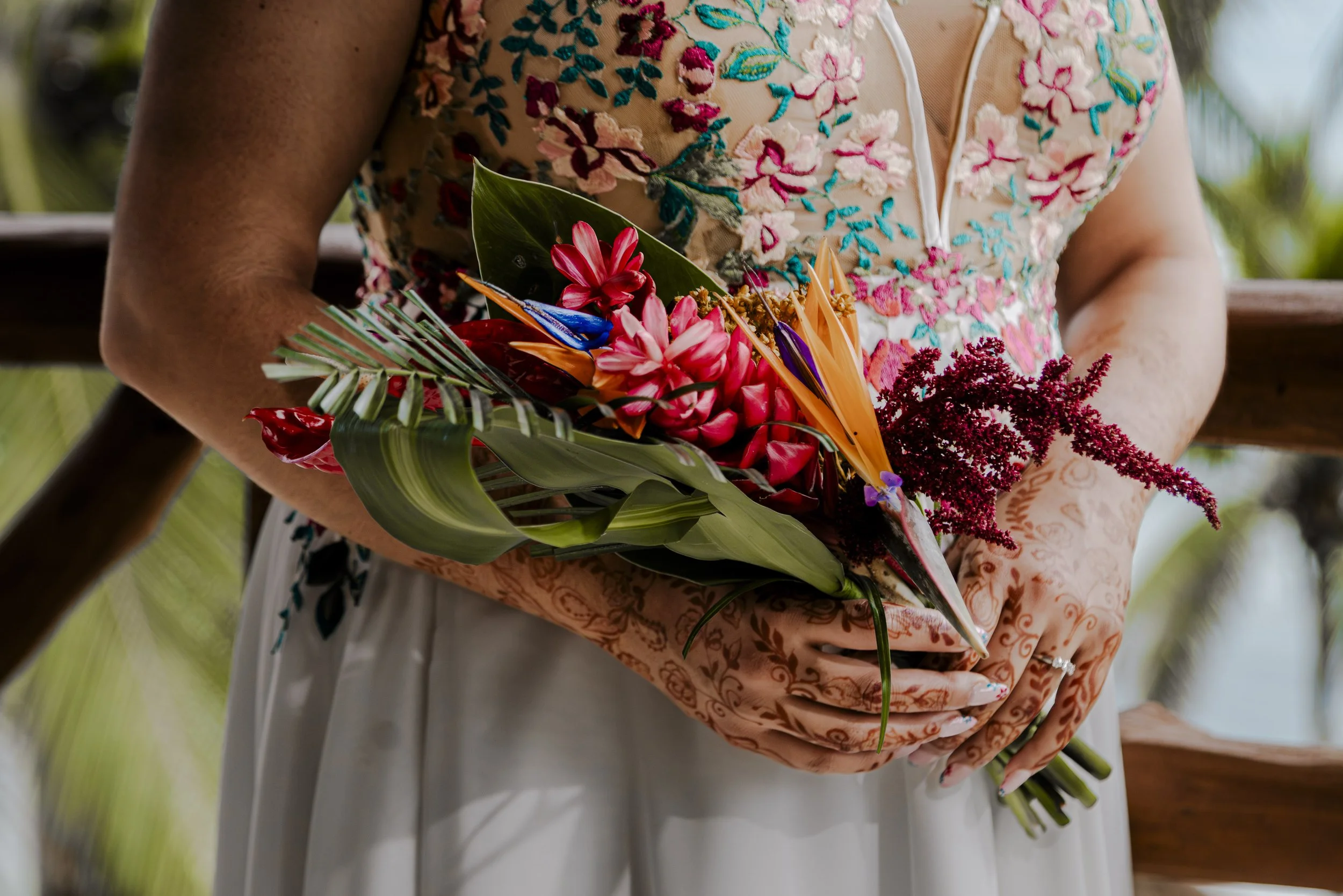 Person wearing a floral embroidered dress holding a bouquet of tropical flowers and green leaves.