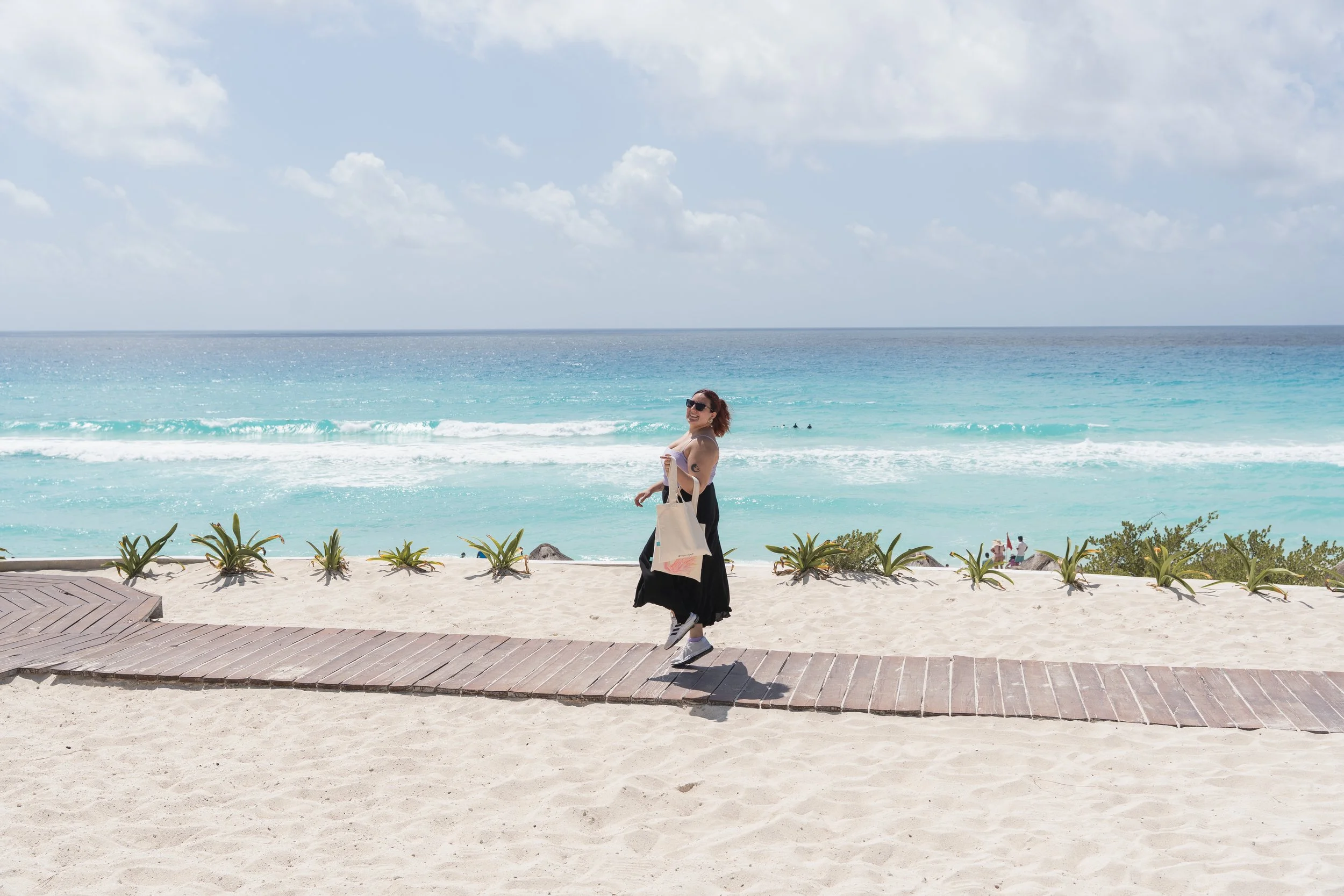 Woman in black dress and sunglasses walking on a wooden path at the beach, carrying a tote bag, with blue ocean and partly cloudy sky in the background.