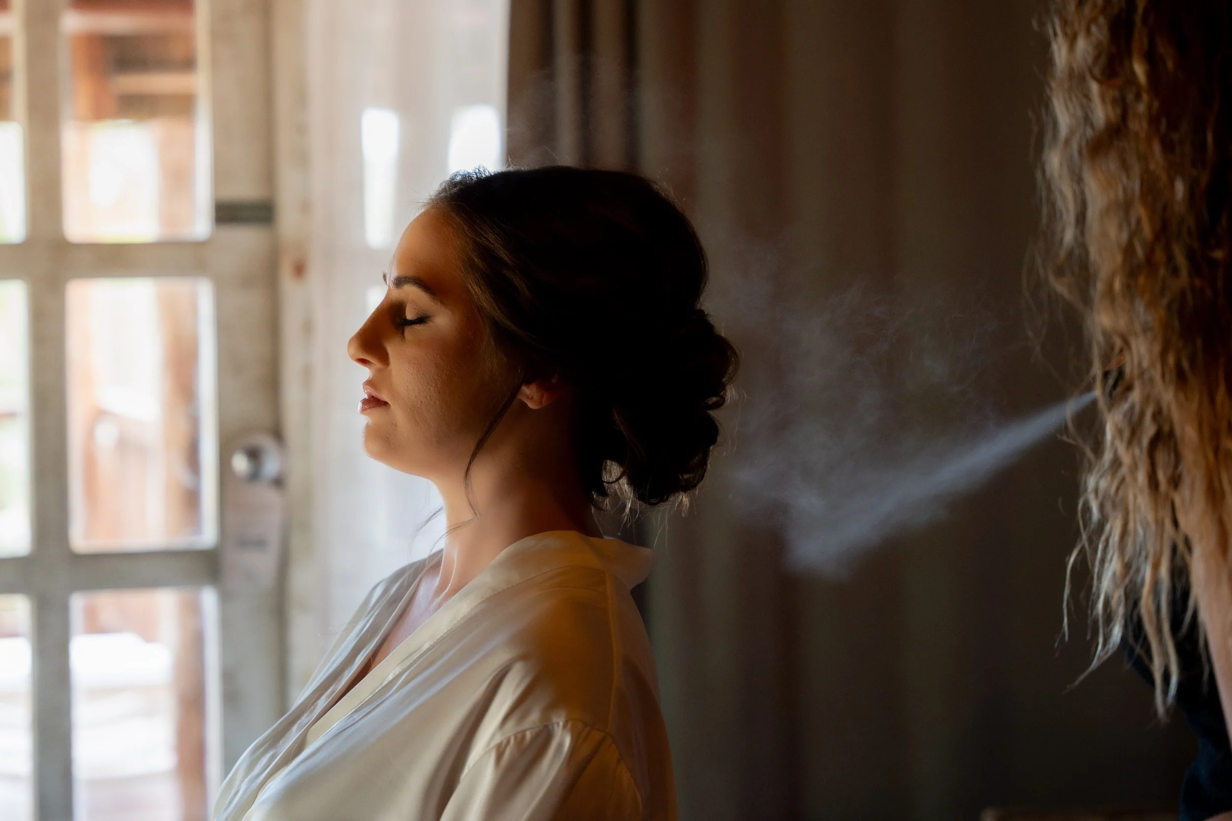 A woman with closed eyes getting her hair sprayed with mist by a stylist.