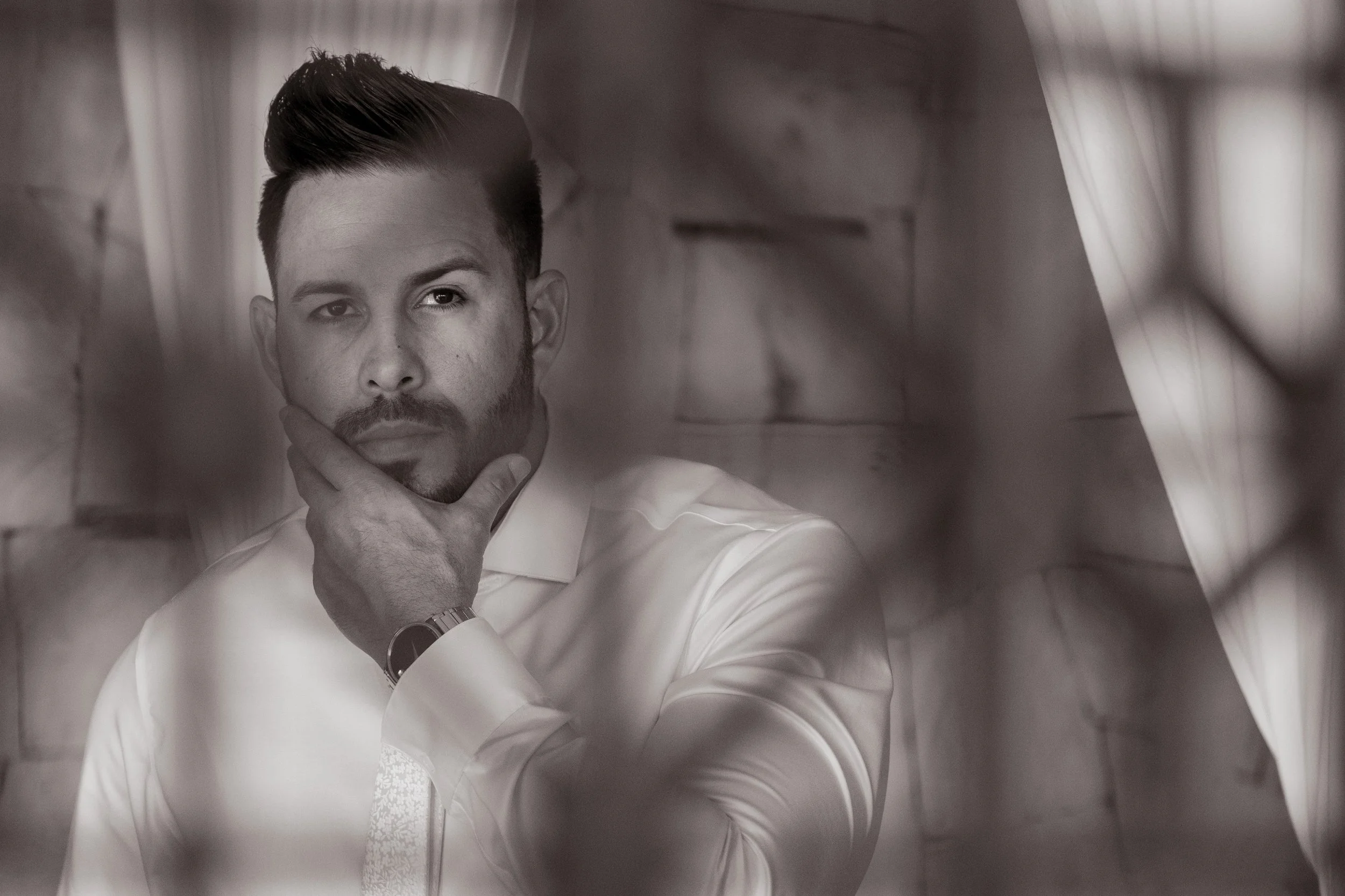 A black and white portrait of a man with styled hair, beard, and mustache, wearing a white shirt, with his hand on his chin, looking at the camera.