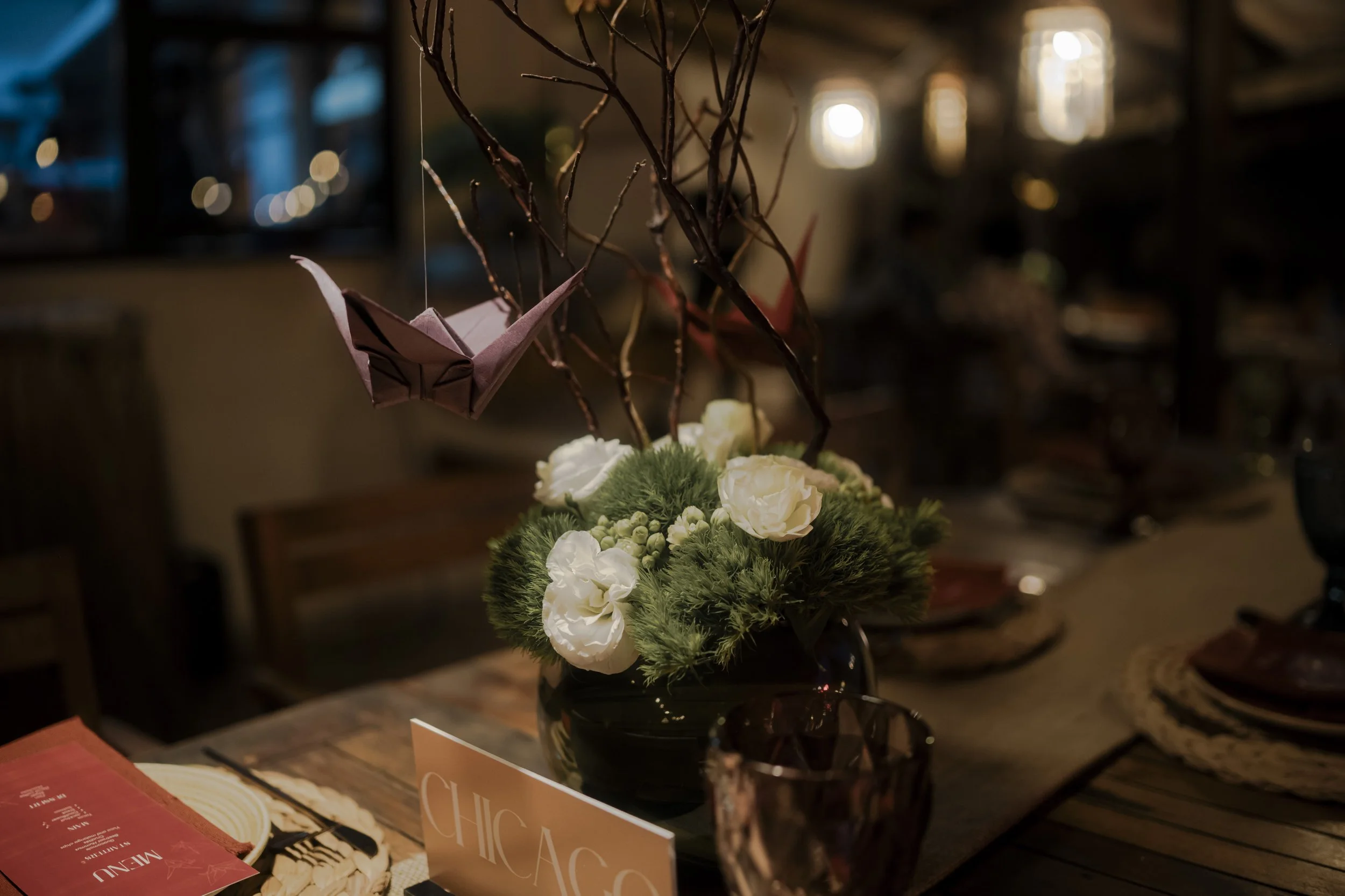 A floral centerpiece with white flowers and greenery on a wooden dining table, with hanging origami cranes, a glass, and a menu card nearby, in a dimly lit restaurant setting.