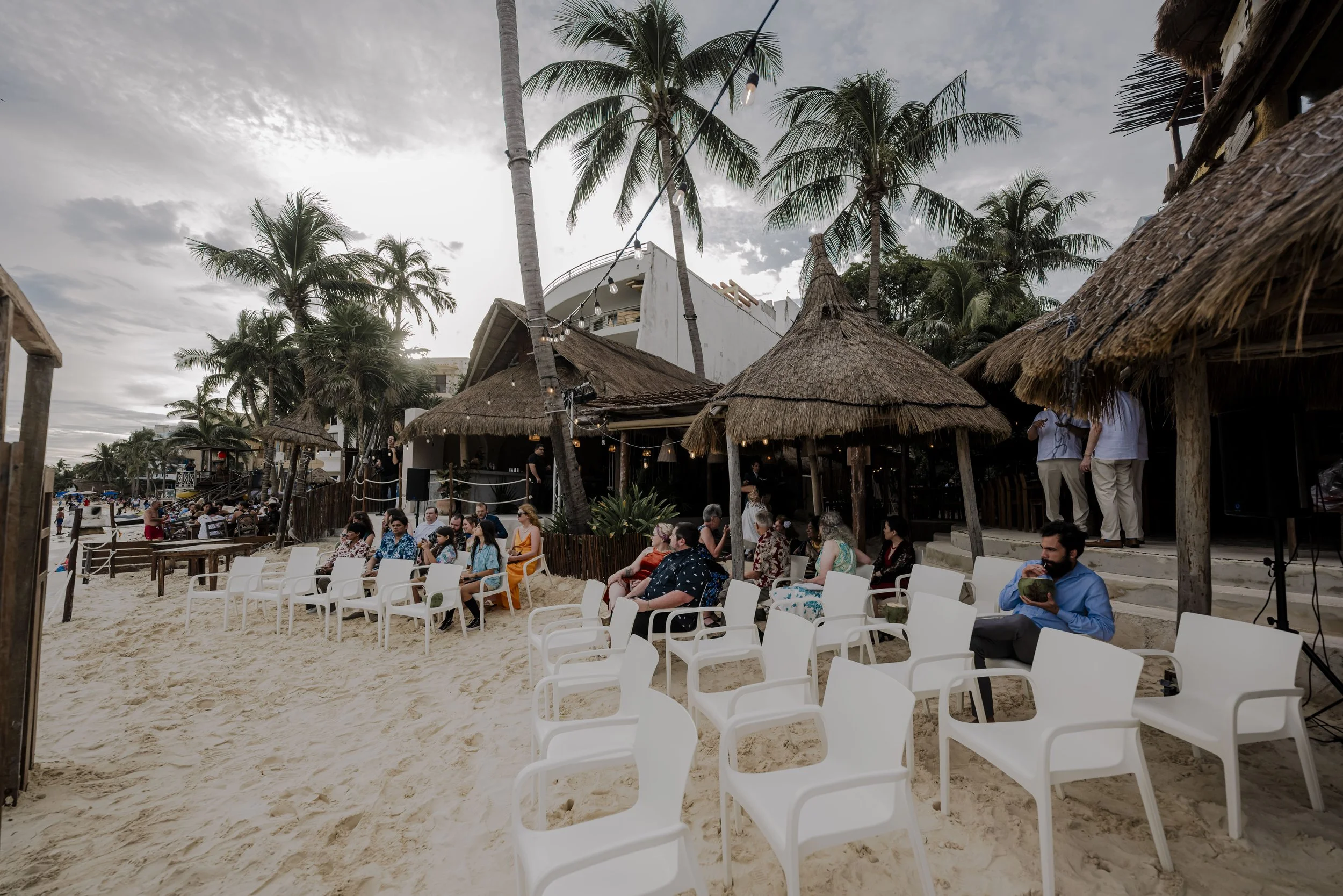 People seated on white plastic chairs on sandy beach, under thatched umbrellas, with palm trees and beachside restaurant in the background.