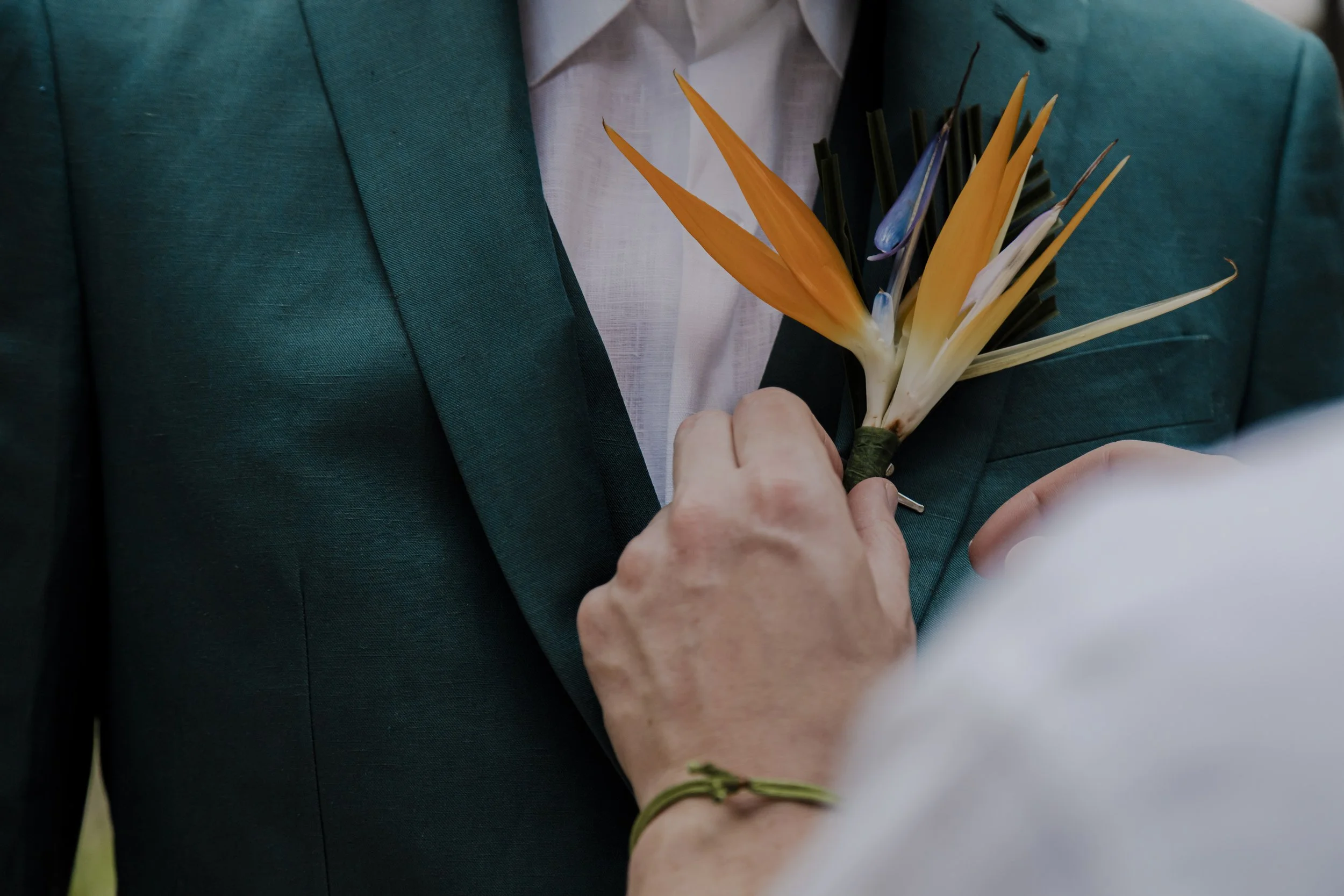 A person in a white shirt is pinning a small flower arrangement, including orange, white, and purple flowers, onto a man's teal suit jacket during a wedding or formal event.
