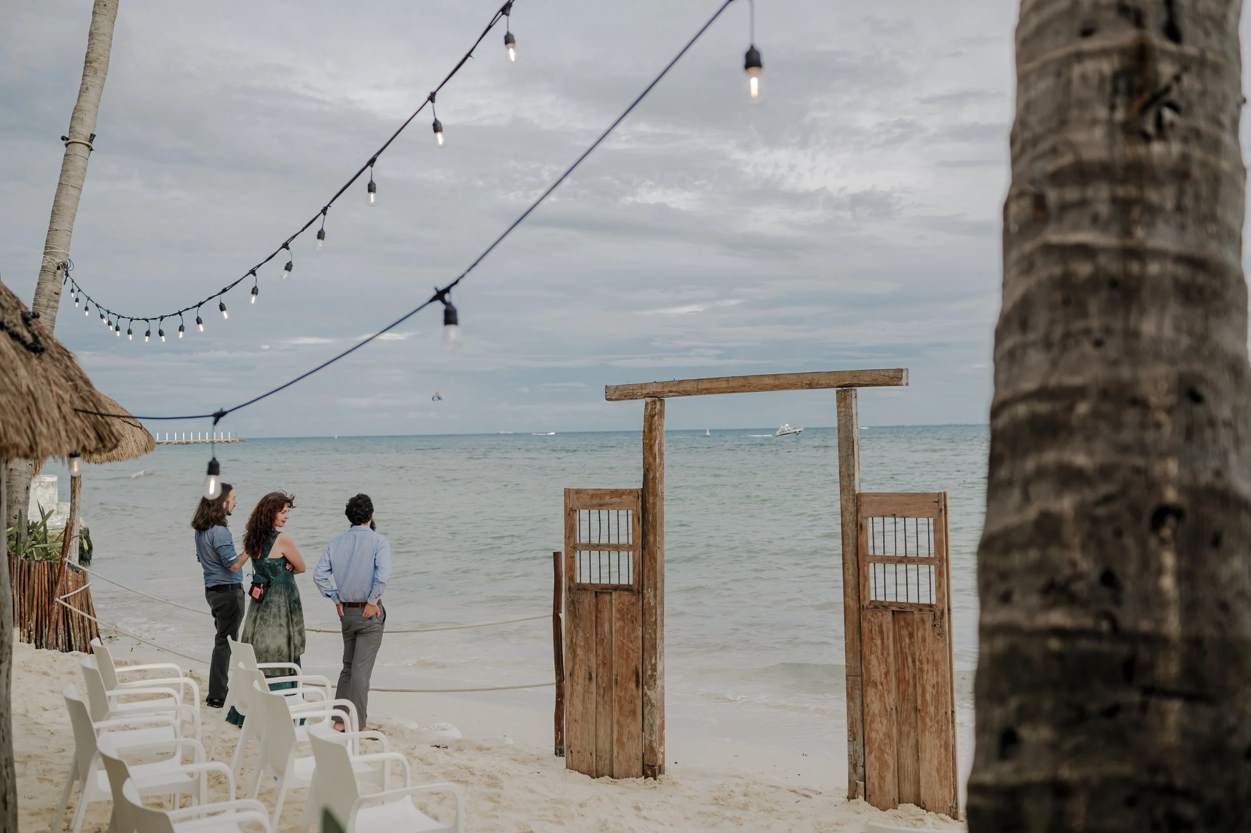 Beachside wedding setup on sandy shore with string lights, chairs, and wooden arch, overlooking ocean with cloudy sky.