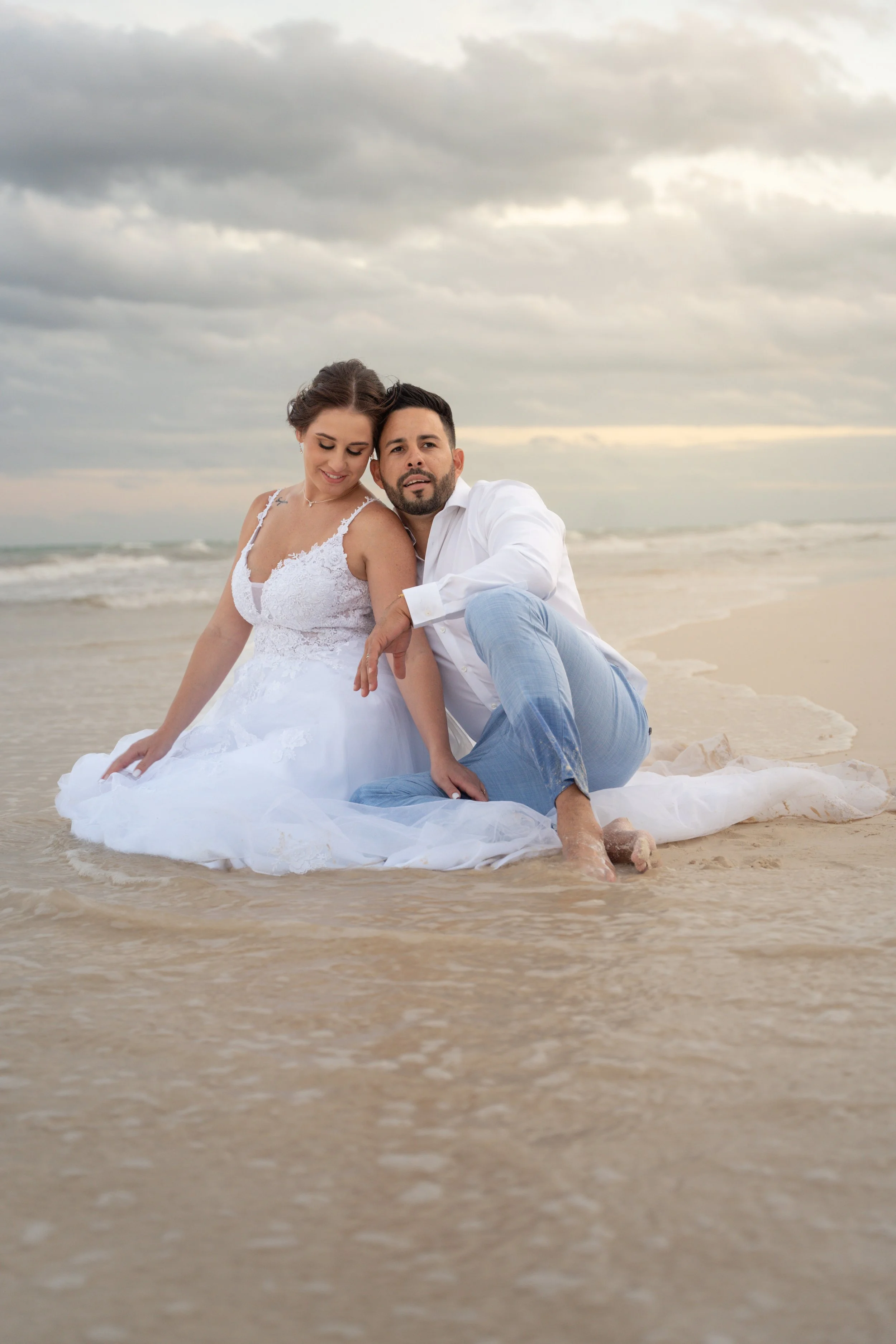 A newlywed couple sitting on the beach, with the bride wearing a white lace wedding gown and the groom in a white shirt and light blue pants, during sunset with cloudy sky.