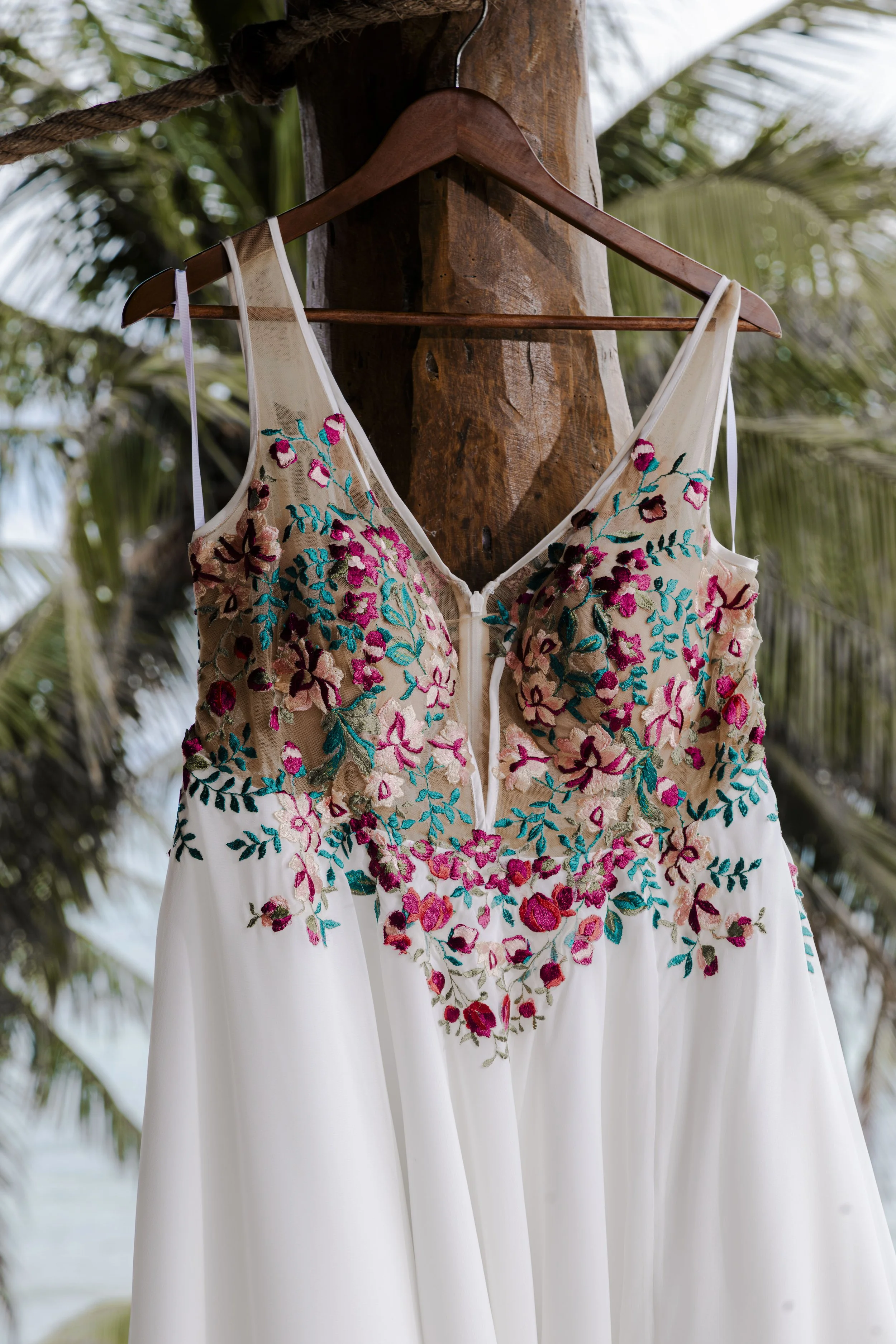A wedding dress with floral embroidery hanging on a wooden hanger against a background of palm trees.