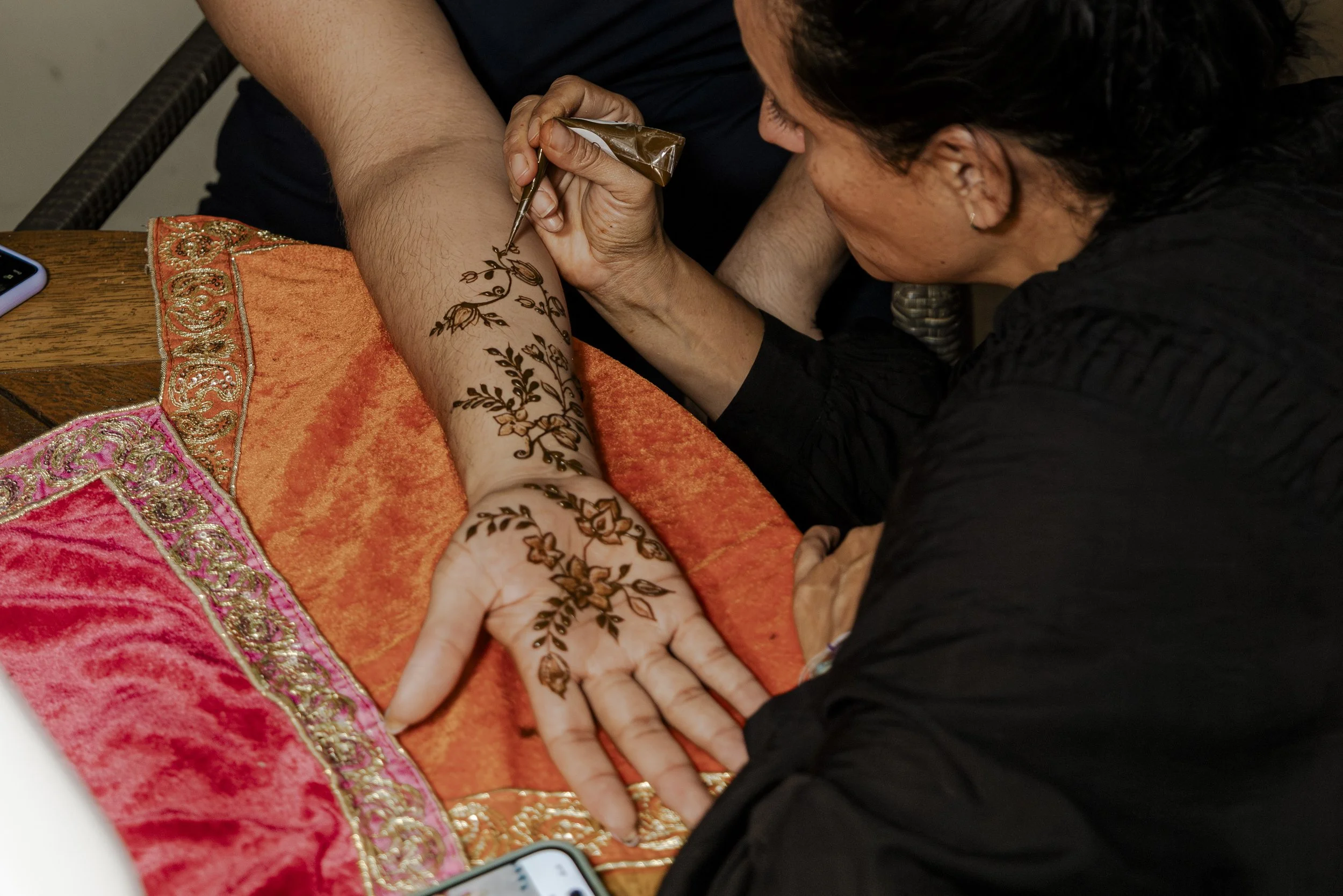 Woman applying mehndi, a traditional henna tattoo, on a person's arm with intricate floral and leaf designs, using a cone. The arm rests on a colorful, embroidered cloth.