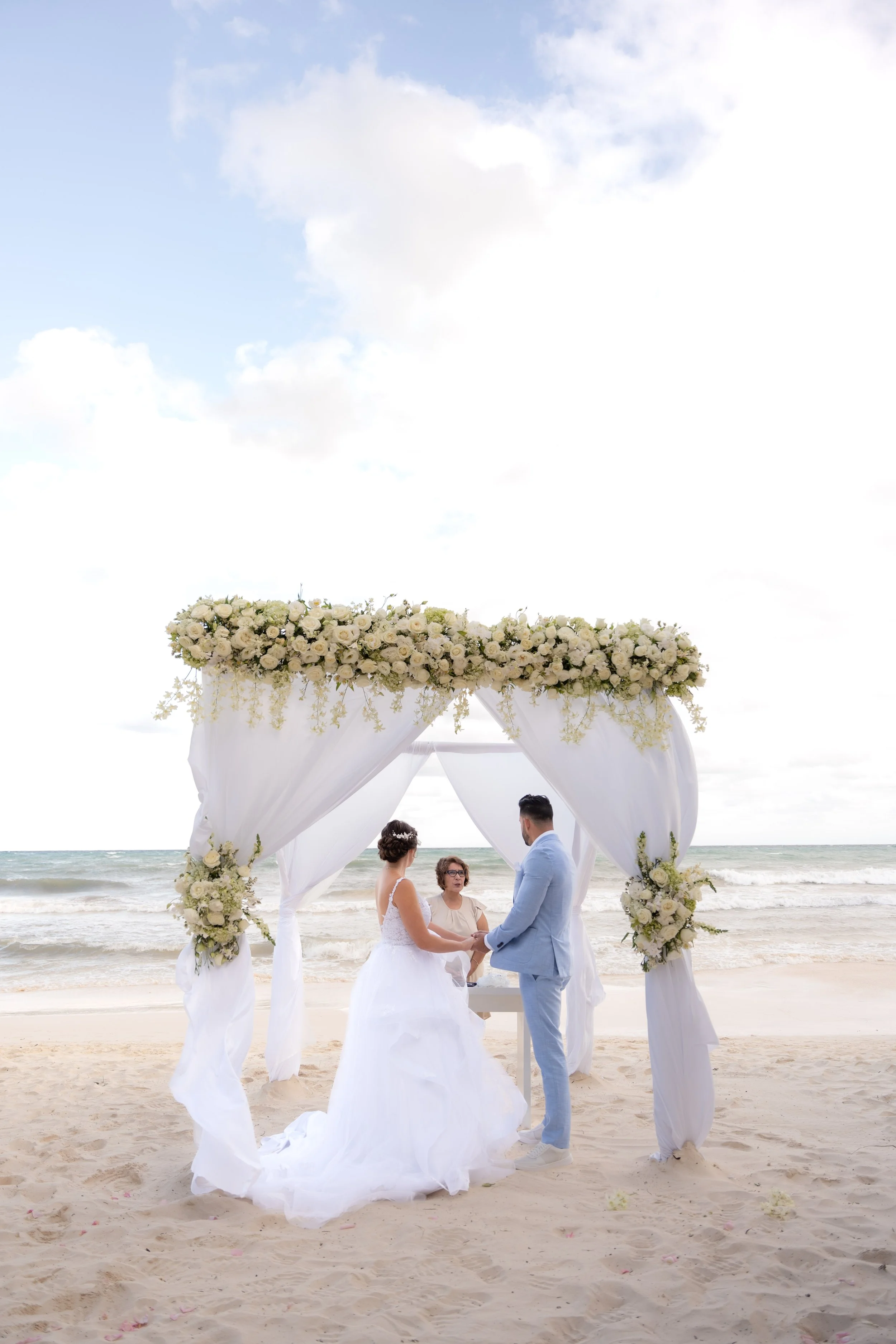 A couple getting married under a beach wedding arch with white flowers and drapery, by the ocean on a sunny day.