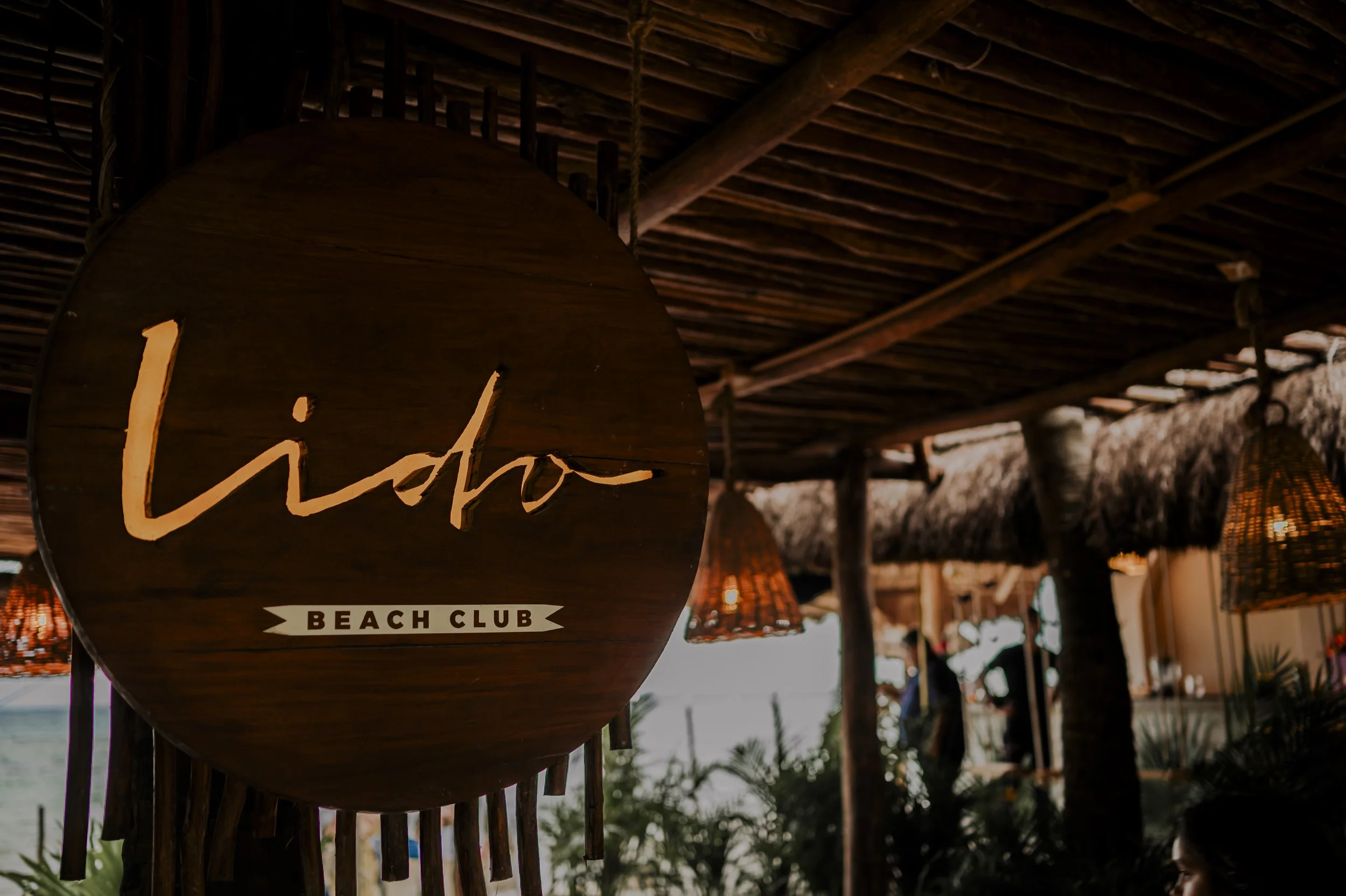 Sign reading 'hido BEACH CLUB' hanging inside a beachside tiki hut with rattan hanging lamps.