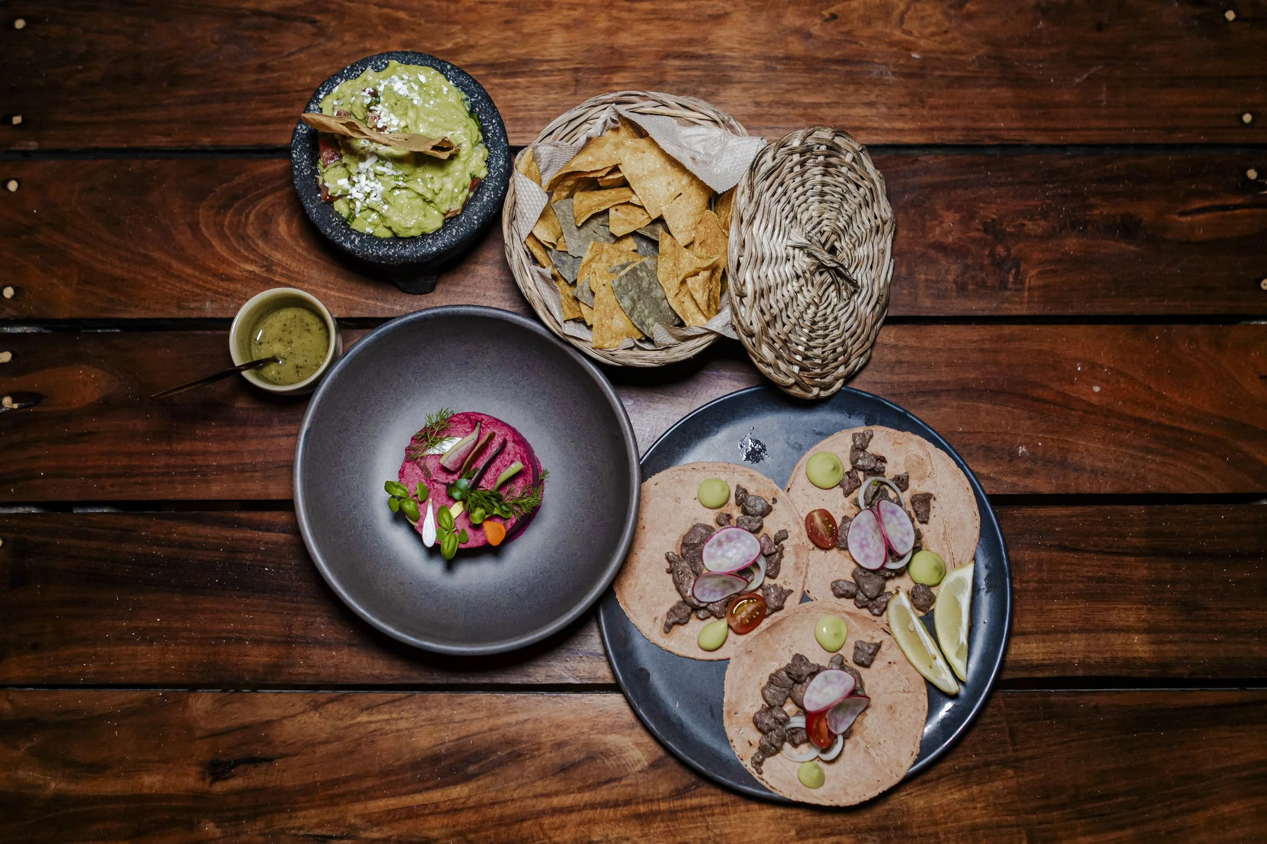 An assortment of Mexican dishes on a wooden table, including three tacos with meat, radish slices, and cherry tomatoes, a bowl of pink beet hummus with herbs, a small bowl of guacamole with a chip, a basket of tortilla chips, and a small container of