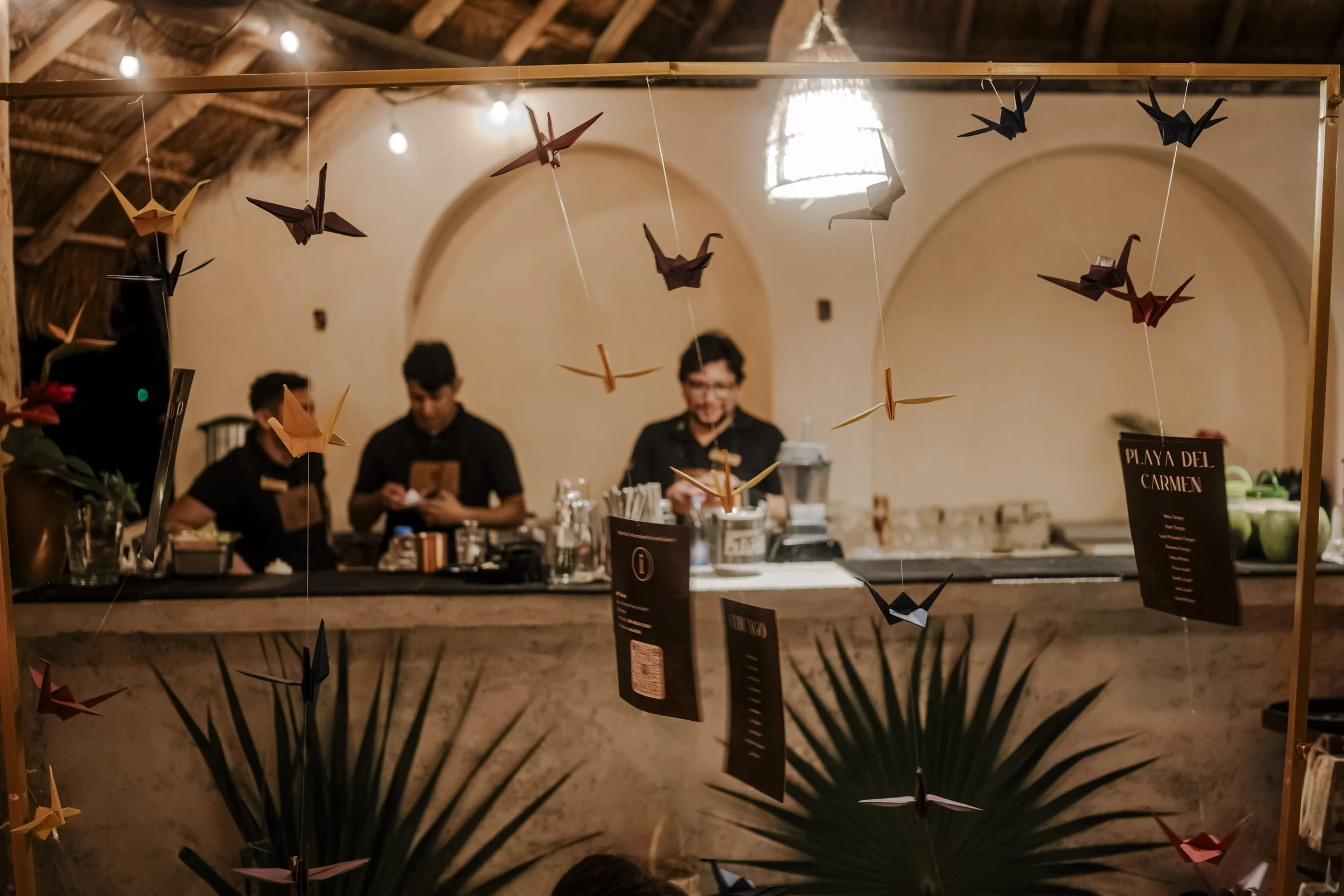 A bar with three bartenders standing behind the counter, decorated with hanging origami cranes and a sign that reads "Playa del Carmen." The background features a warm, rustic interior with arched wall openings and wooden beams.