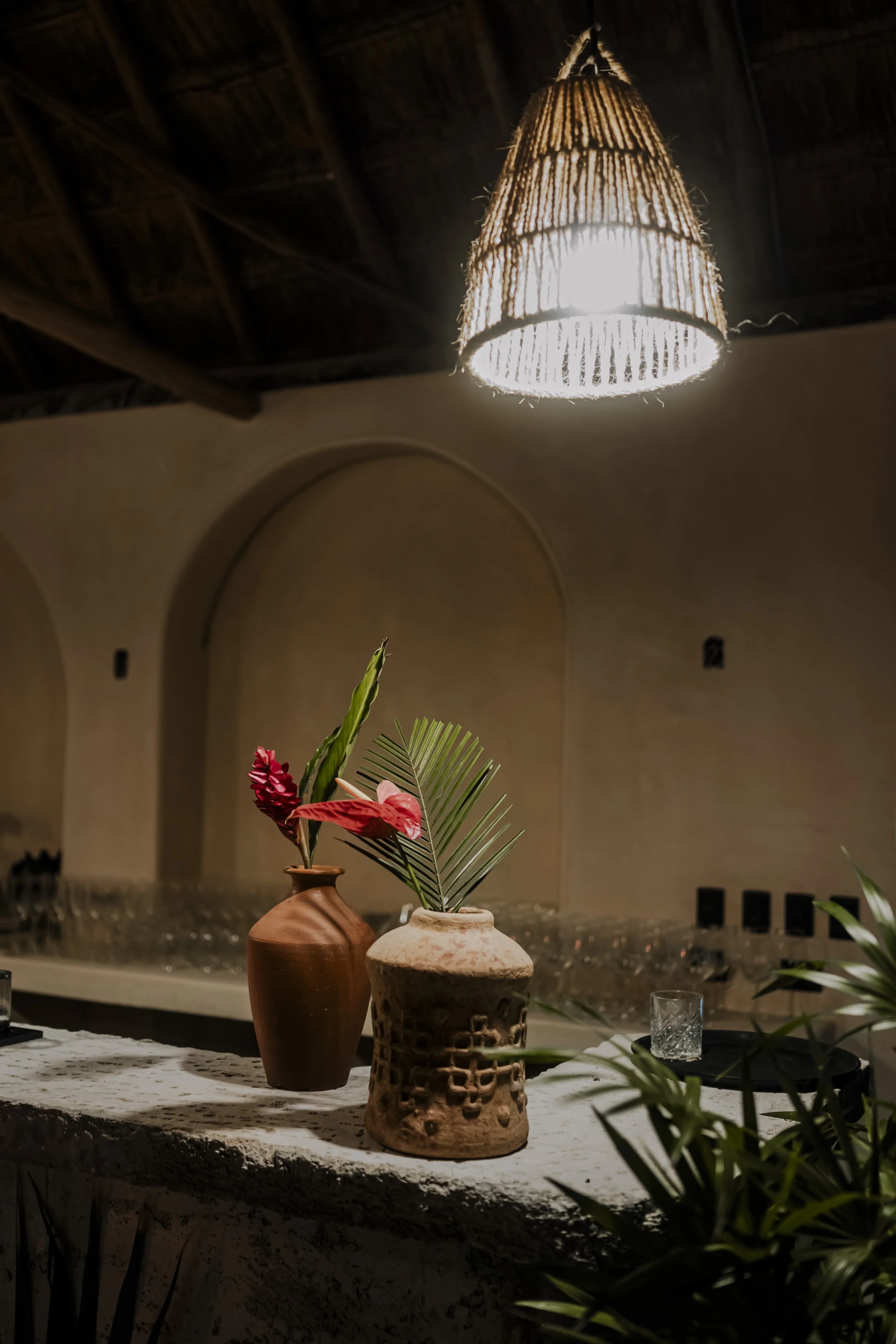 Two vases with pink and green tropical plants on a rustic white bar counter, illuminated by a woven pendant light above in a warm-toned room.
