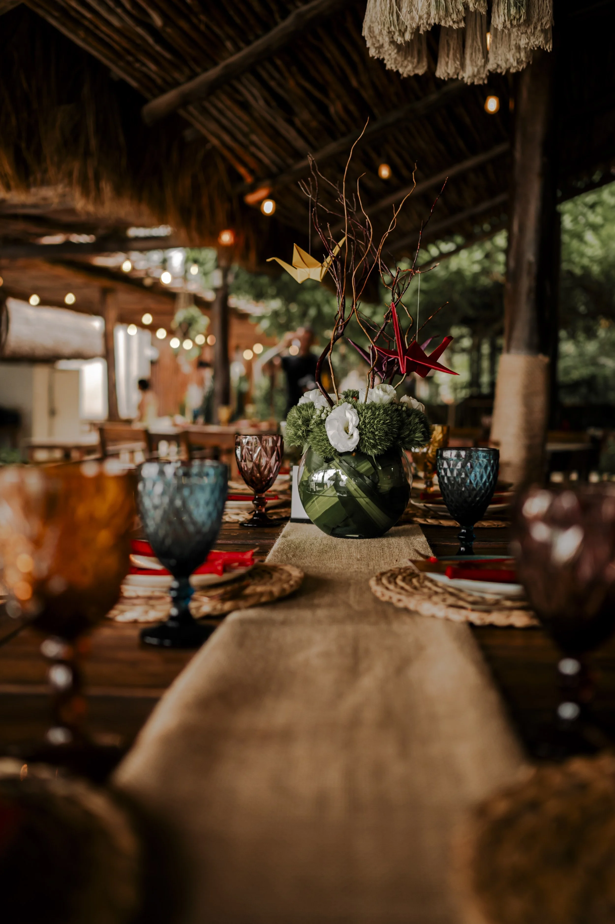 A rustic outdoor dining table with a beige table runner, colorful textured glassware, and a floral centerpiece with white flowers and decorative branches