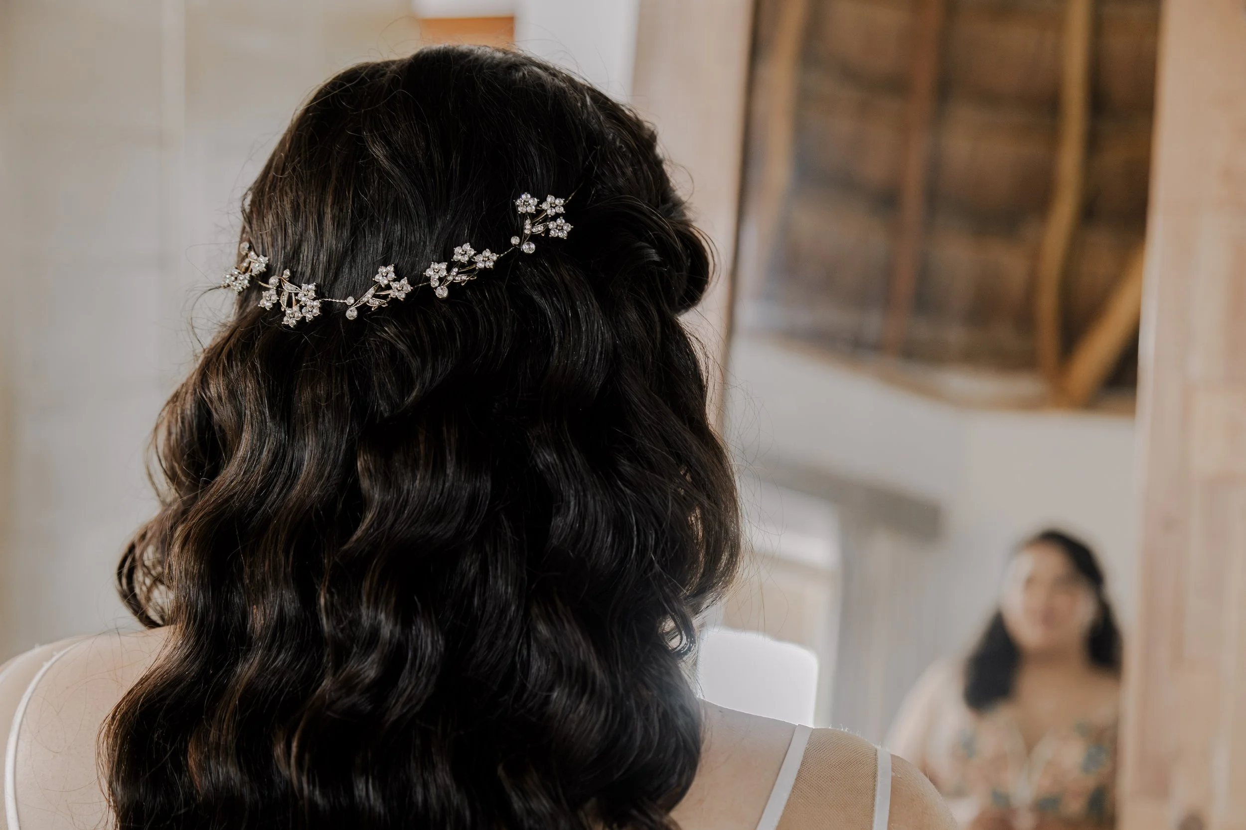 Back view of a woman with dark, wavy hair wearing a floral tiara, looking into a mirror, with another woman visible in the reflection.