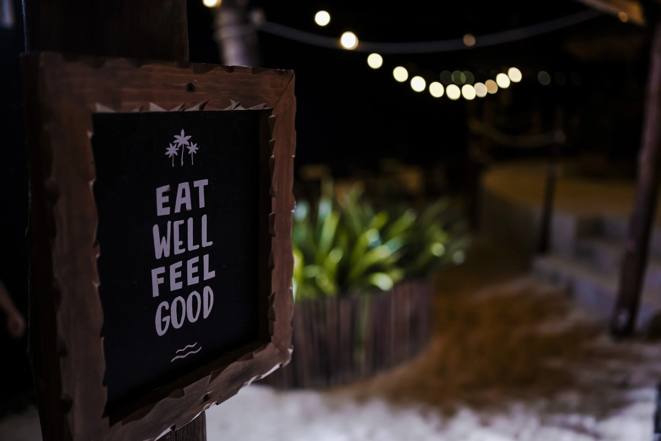 A sign that reads "Eat Well Feel Good" in white letters on a black background, mounted on a wooden frame, with string lights overhead and greenery in the background, at night.