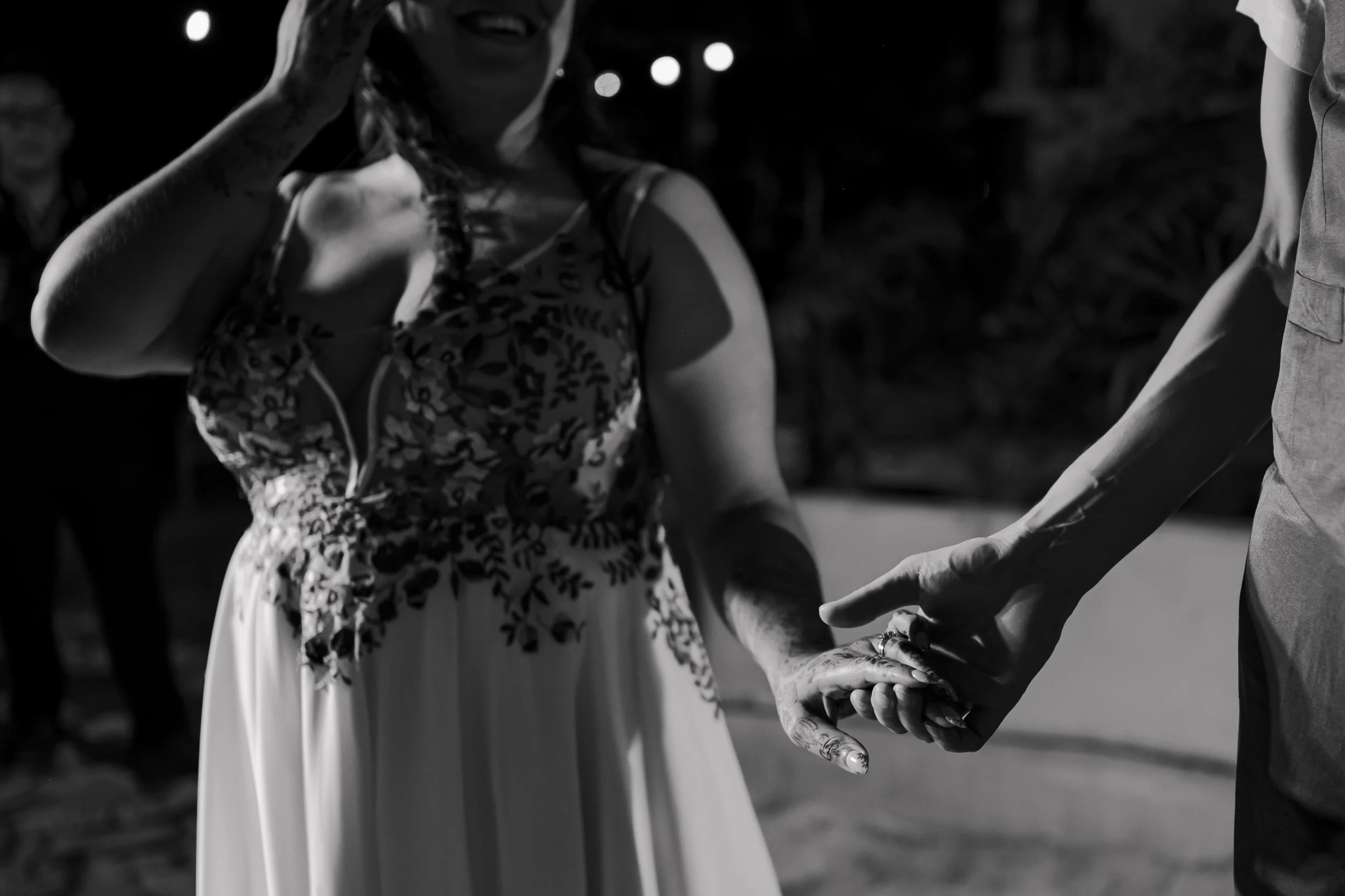 A woman in a floral dress holding hands with a man during a dance at night, with people and lights in the background.