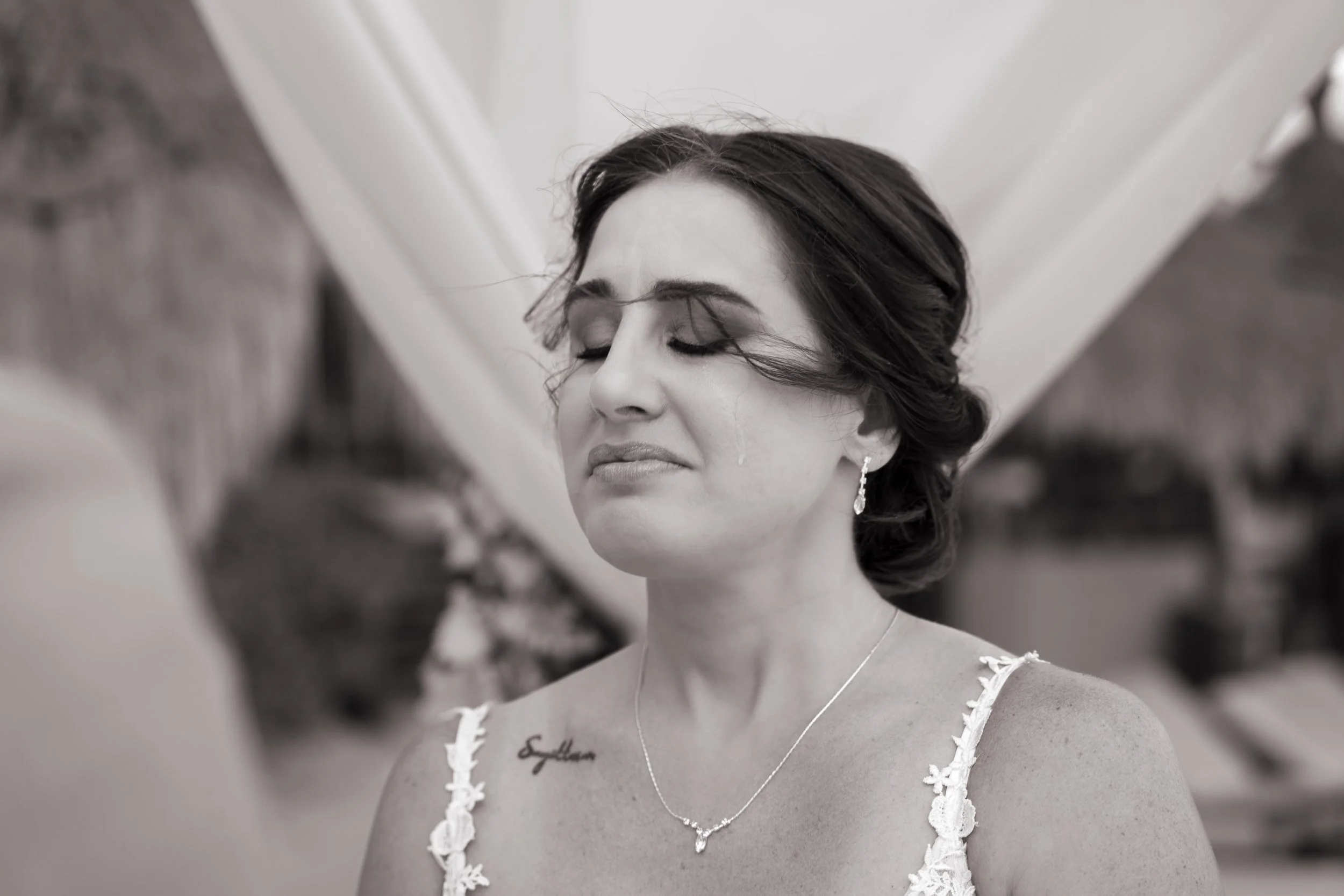 A woman with dark hair styled in an updo, wearing a floral lace dress, earrings, a necklace, and a tattoo on her shoulder, appears emotional and is crying with her eyes closed.