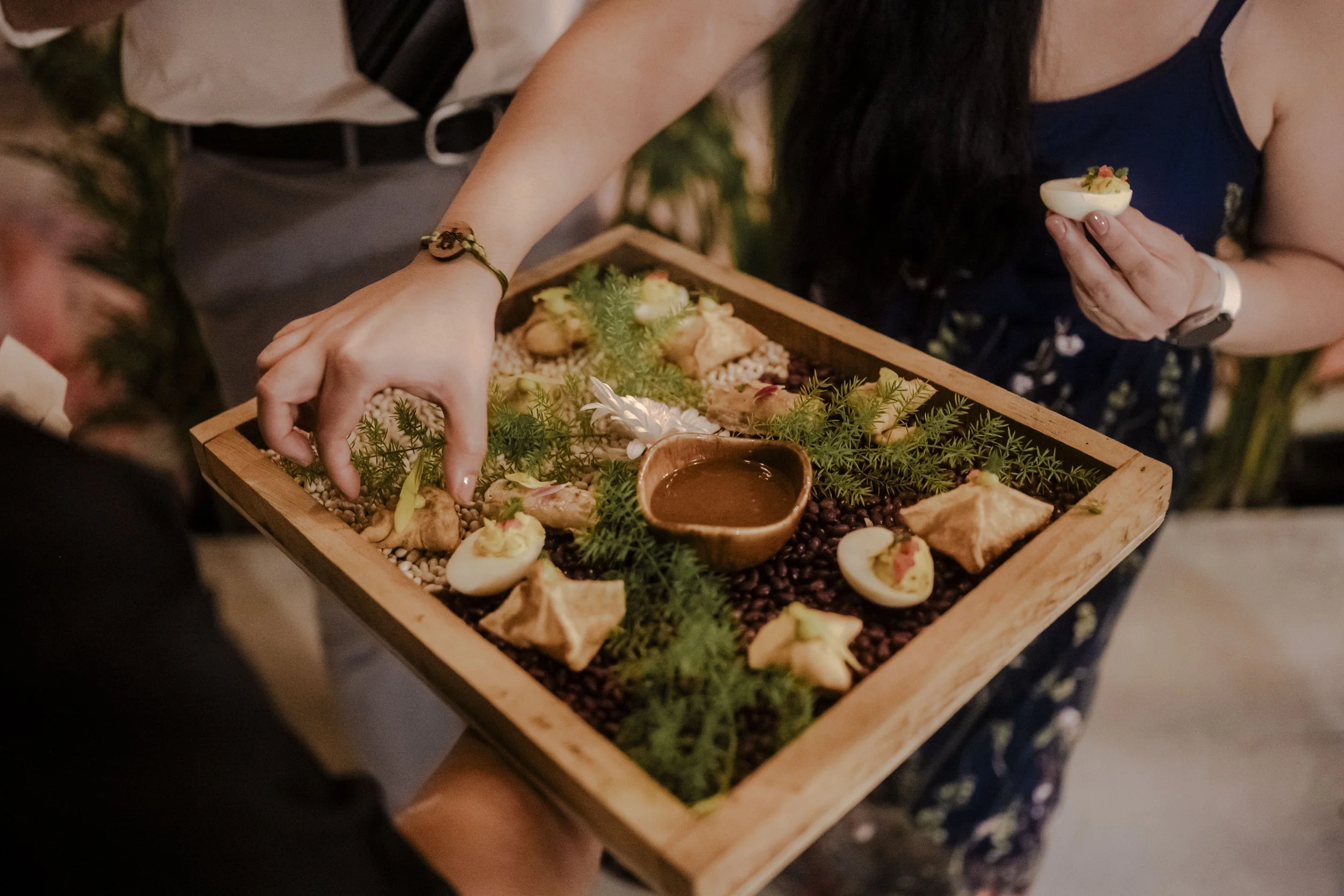 A person holding a wooden tray with appetizers, including deviled eggs, samosas, herbs, and a small bowl of dipping sauce, during a social gathering.