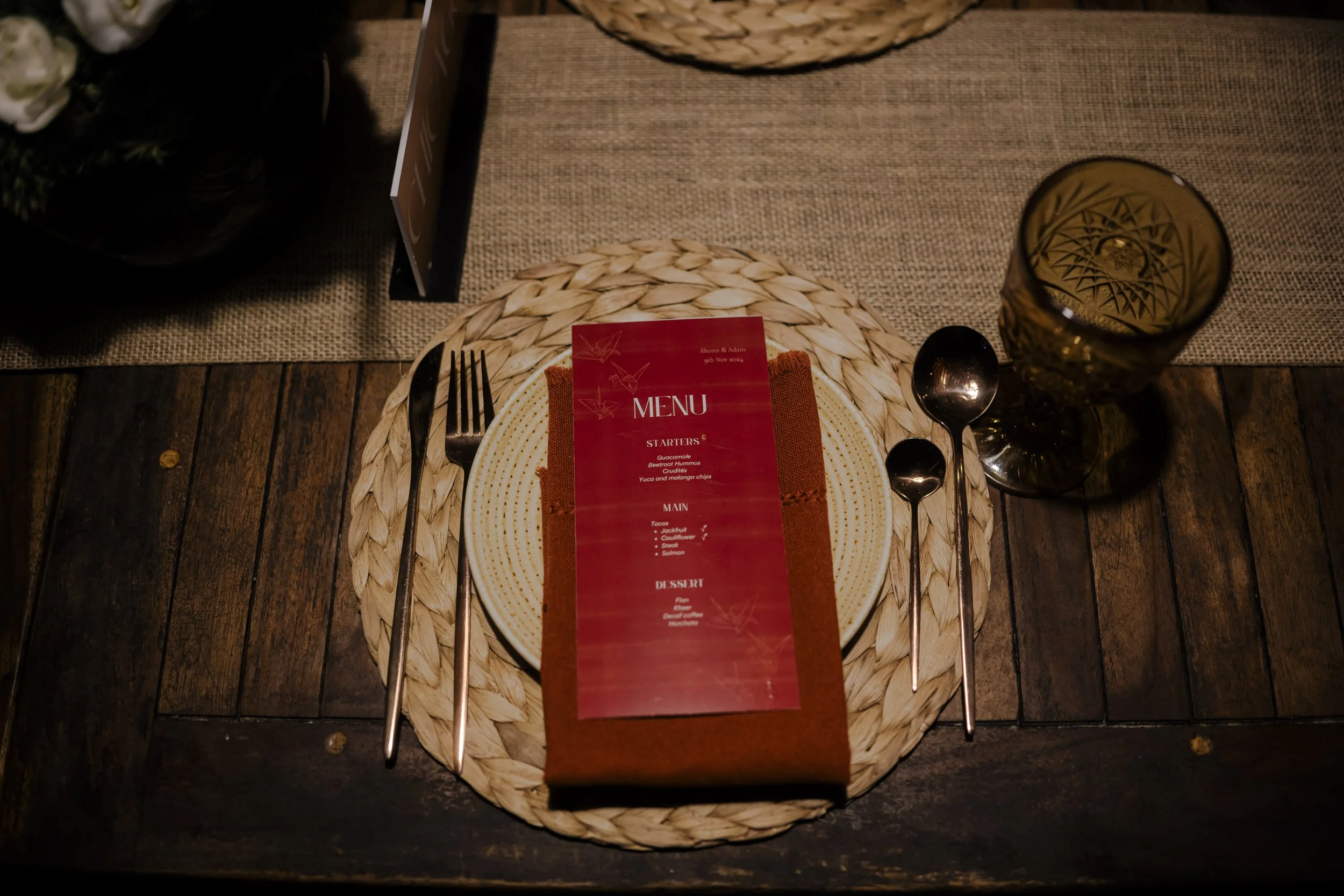 A table setting with a woven placemat, a decorative plate, a folded red napkin, a menu, and silverware, with a glass of water to the right.