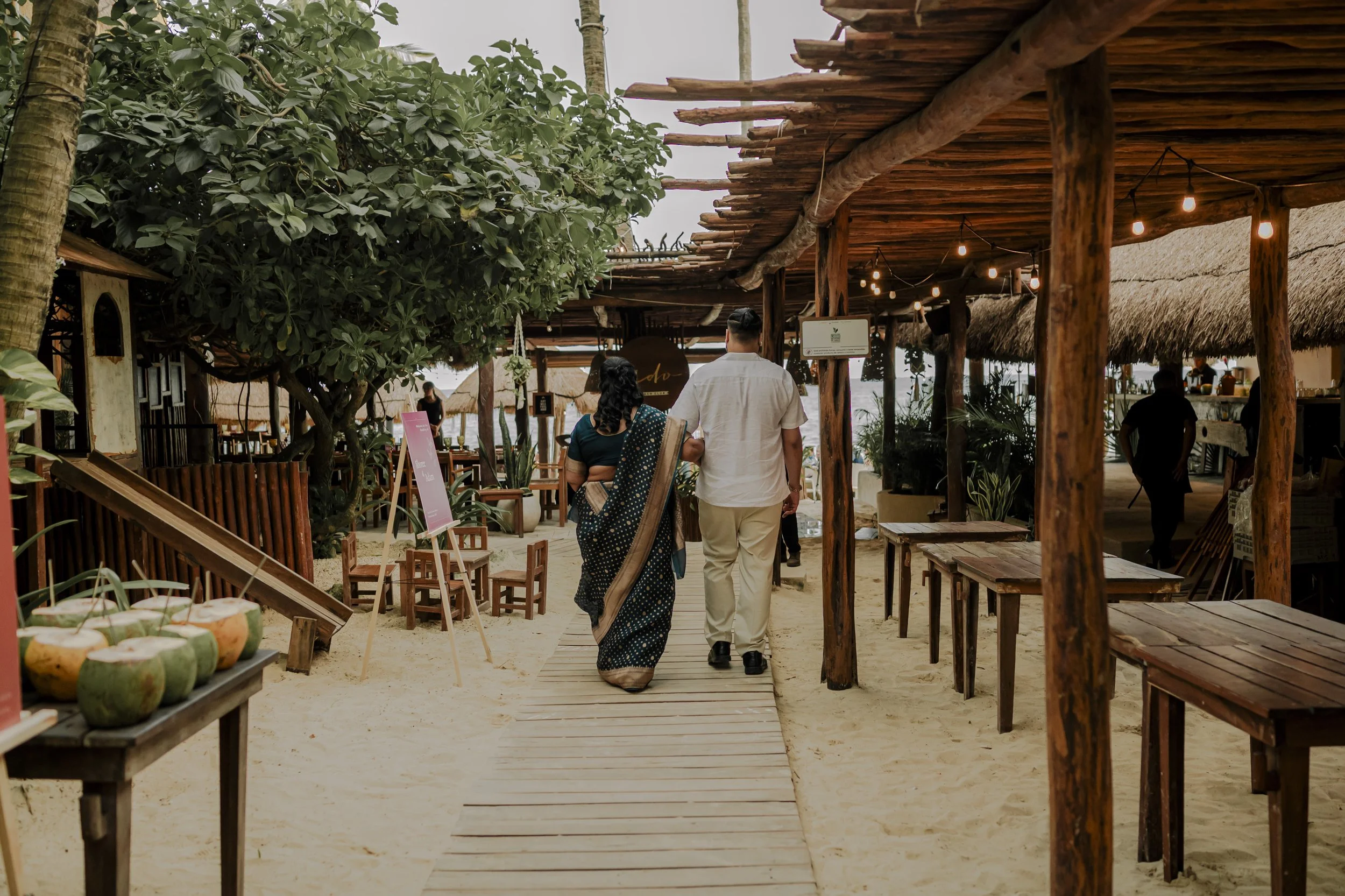 Man and woman walking on a sandy pathway through an outdoor beachside restaurant, with wooden tables, stools, and string lights, surrounded by lush green trees and thatched-roof structures.
