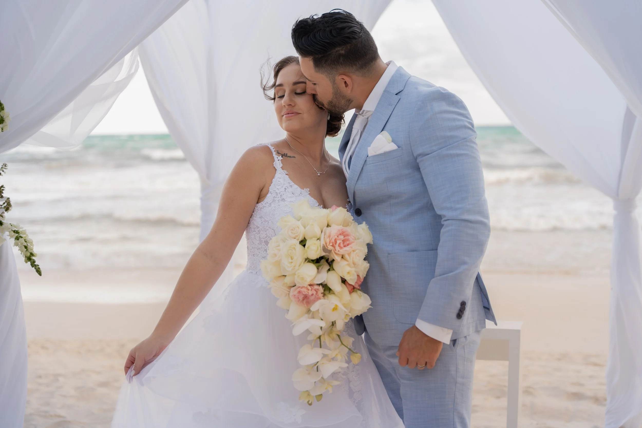 A bride and groom sharing a kiss under a white wedding arch on the beach, with the ocean in the background.