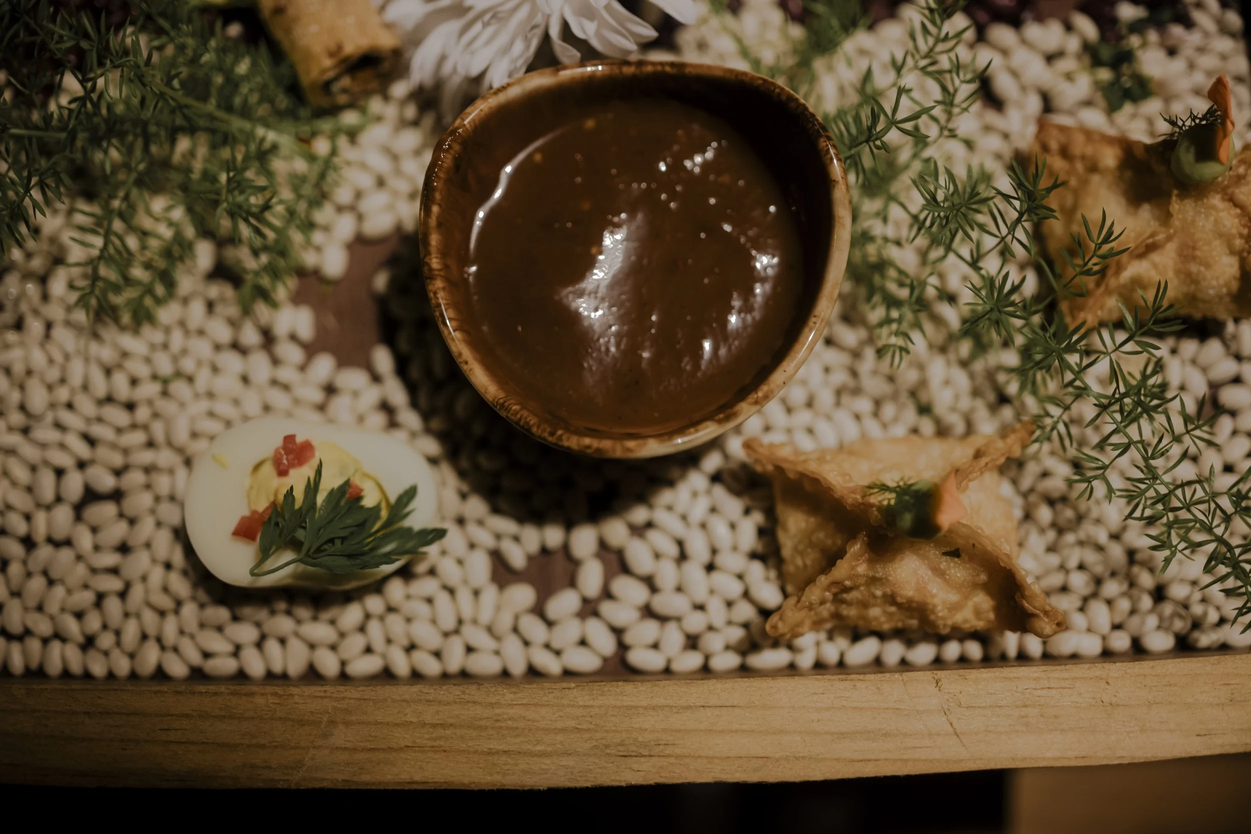 Appetizer plate with a small bowl of brown sauce, boiled egg garnished with greenery and red toppings, and fried wontons garnished with herbs on a bed of white beans, surrounded by green herbs.