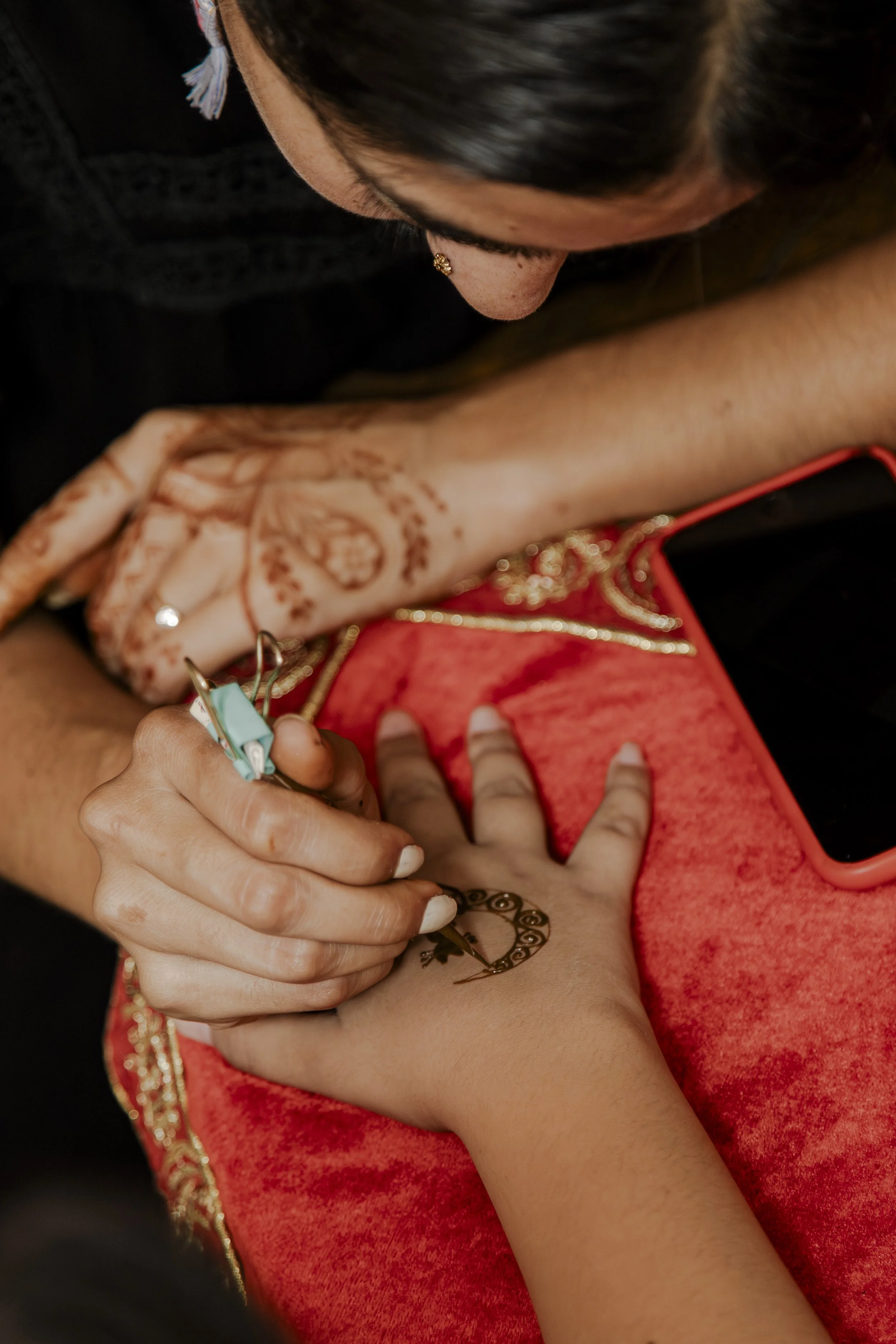 A woman is getting a henna tattoo on her hand. The woman, dressed in a red and gold outfit, is seated on a red cloth. She has henna designs on her hand, and the artist is carefully applying the design with a small tool. A smartphone with a red case i