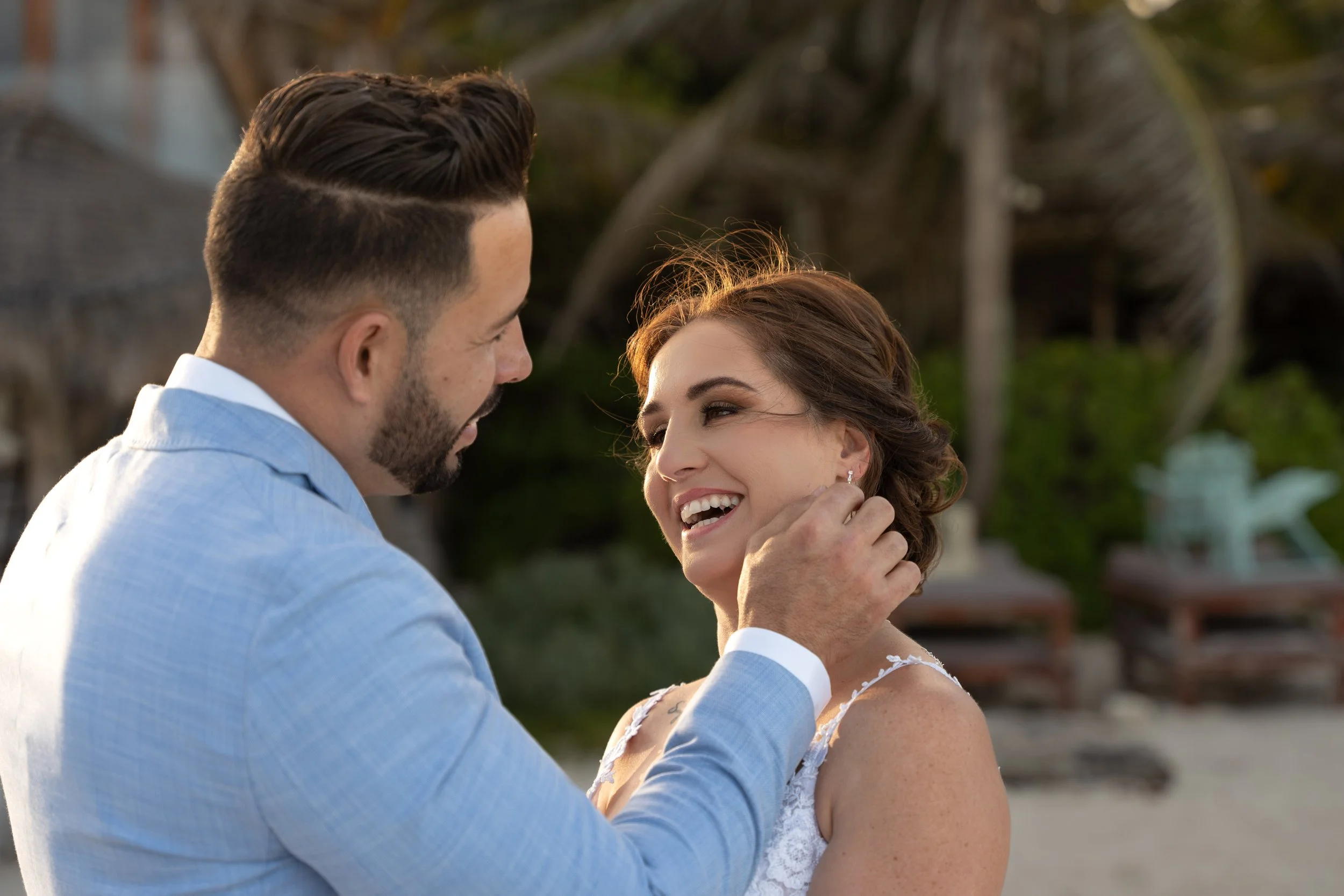 A man gently touching a woman's face while smiling at her during a romantic moment on a beach, with palm trees and outdoor furniture in the background.