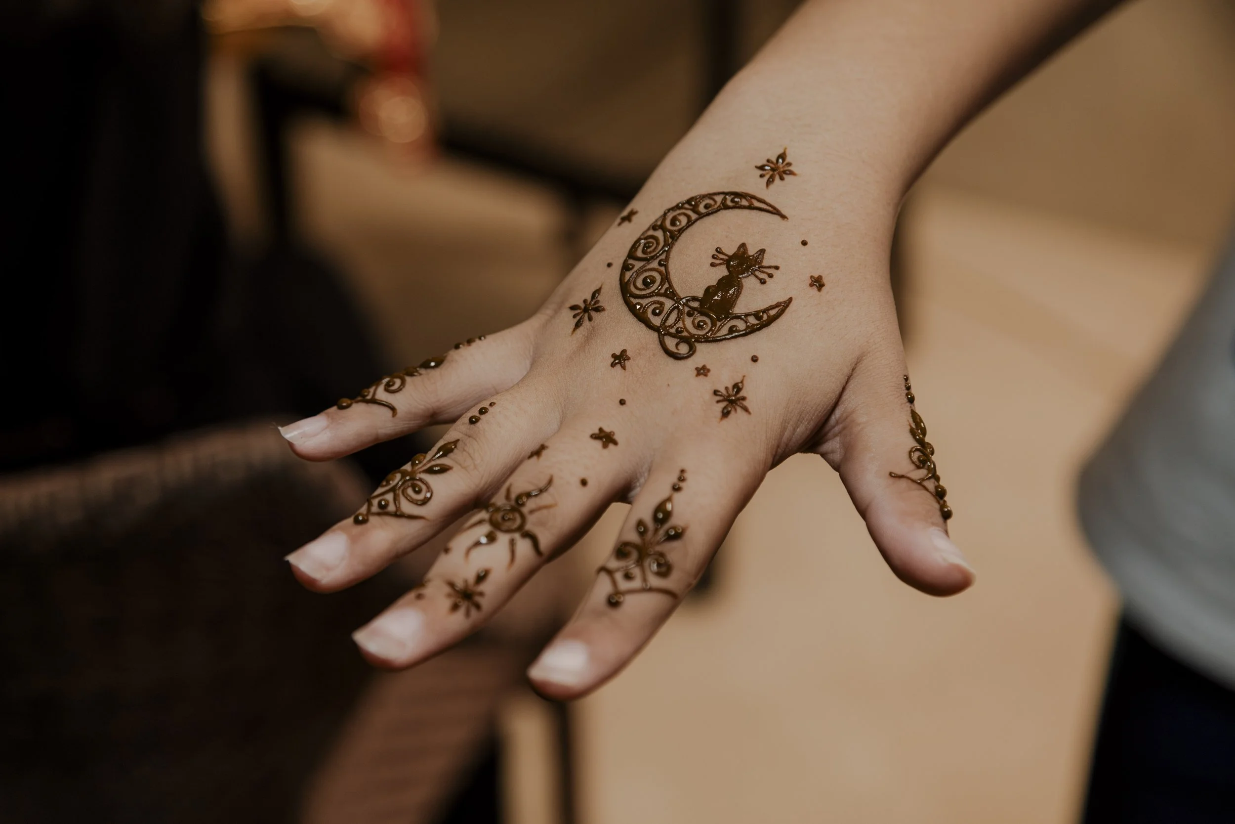 Hand with henna designs featuring a crescent moon, cat silhouette, stars, and floral patterns.