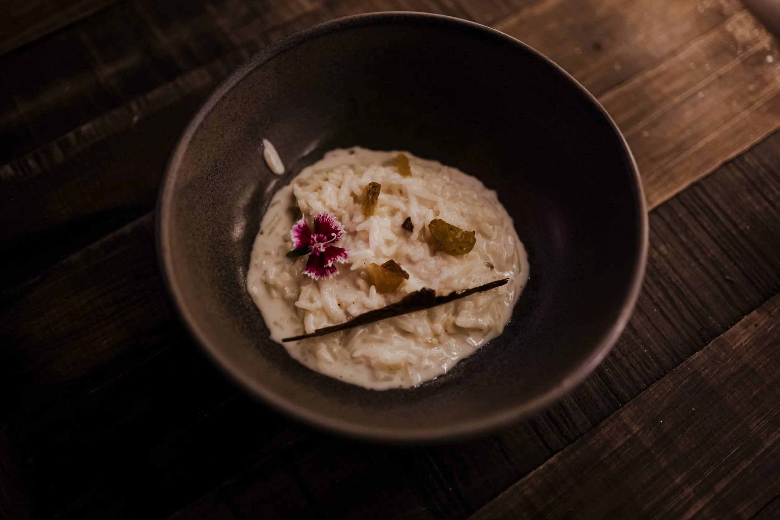 A bowl of creamy risotto garnished with an edible flower and small herbs, served on a dark wooden table.