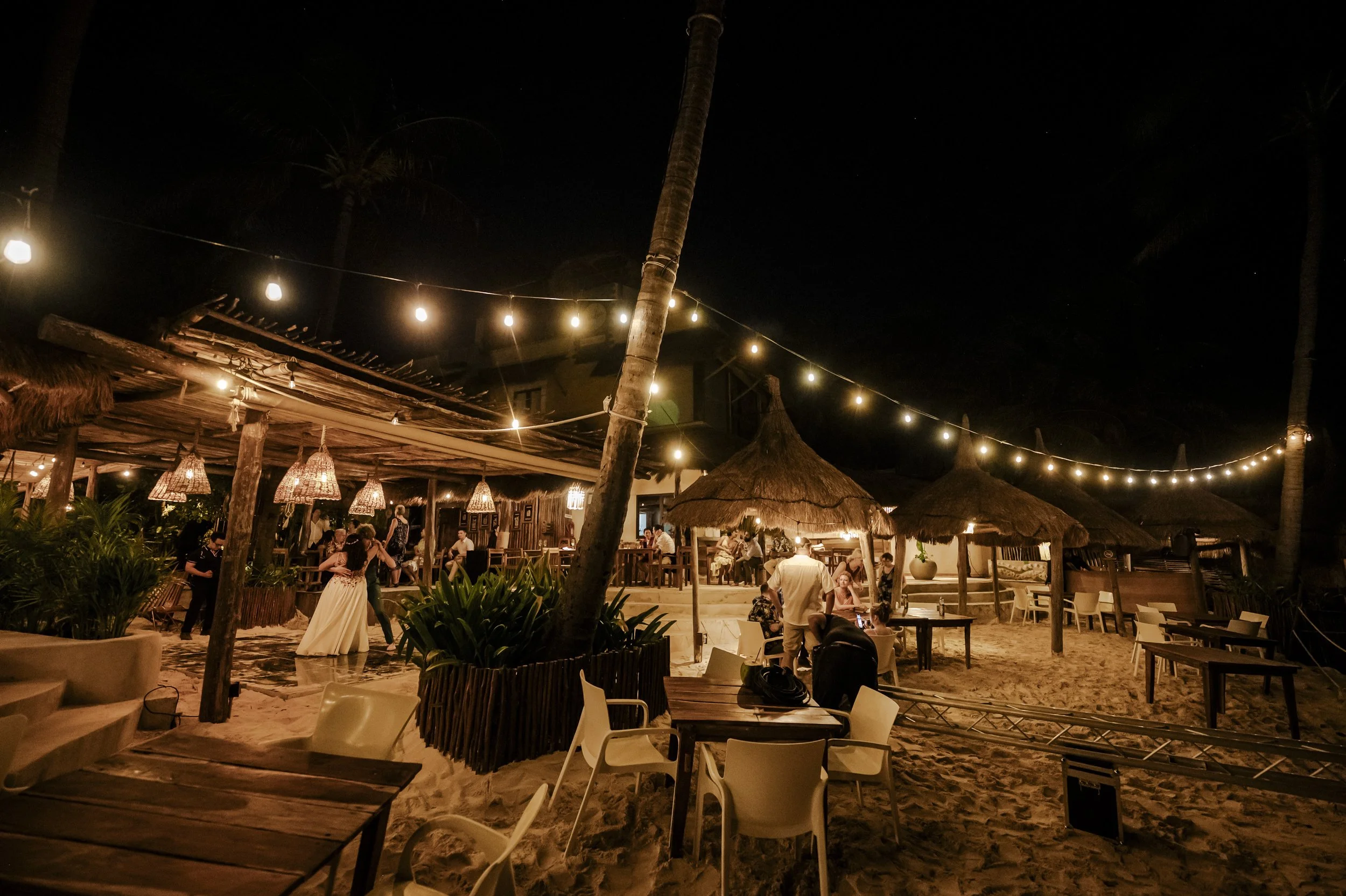 Nighttime beach scene with string lights, a thatched-roof tiki hut, people dining and dancing, sandy ground, and tropical decor.