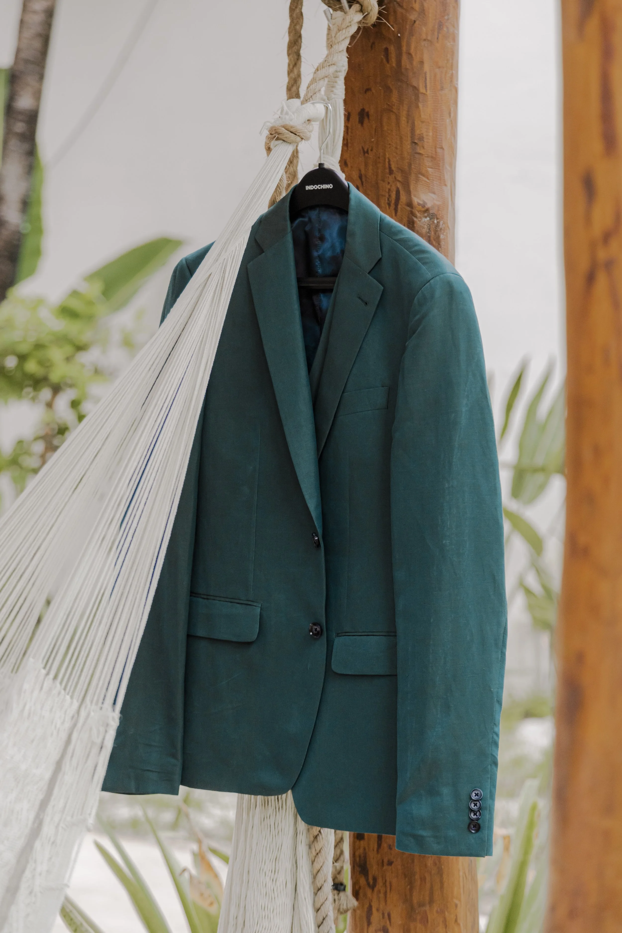 Teal men's suit jacket hanging on a hanger attached to a wooden pole, with a hammock and tropical plants in the background.