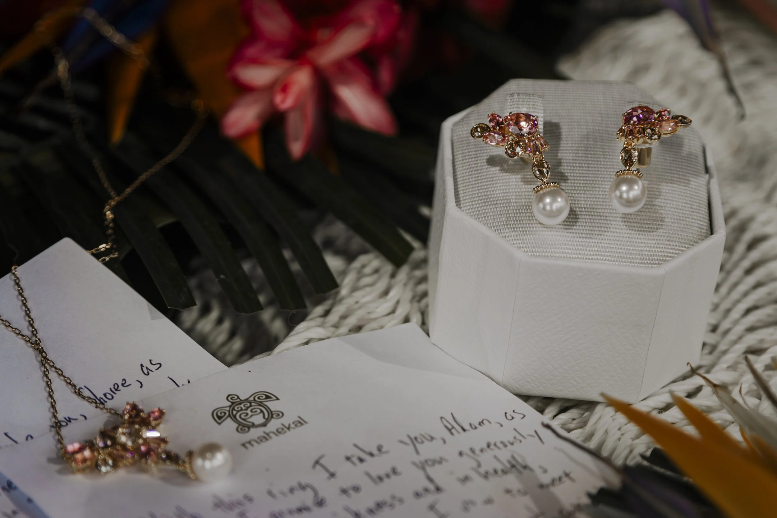 A pair of pink and gold earrings with pearl drops displayed on a white jewelry box, a gold necklace with a pendant, a handwritten letter, and flowers in the background.
