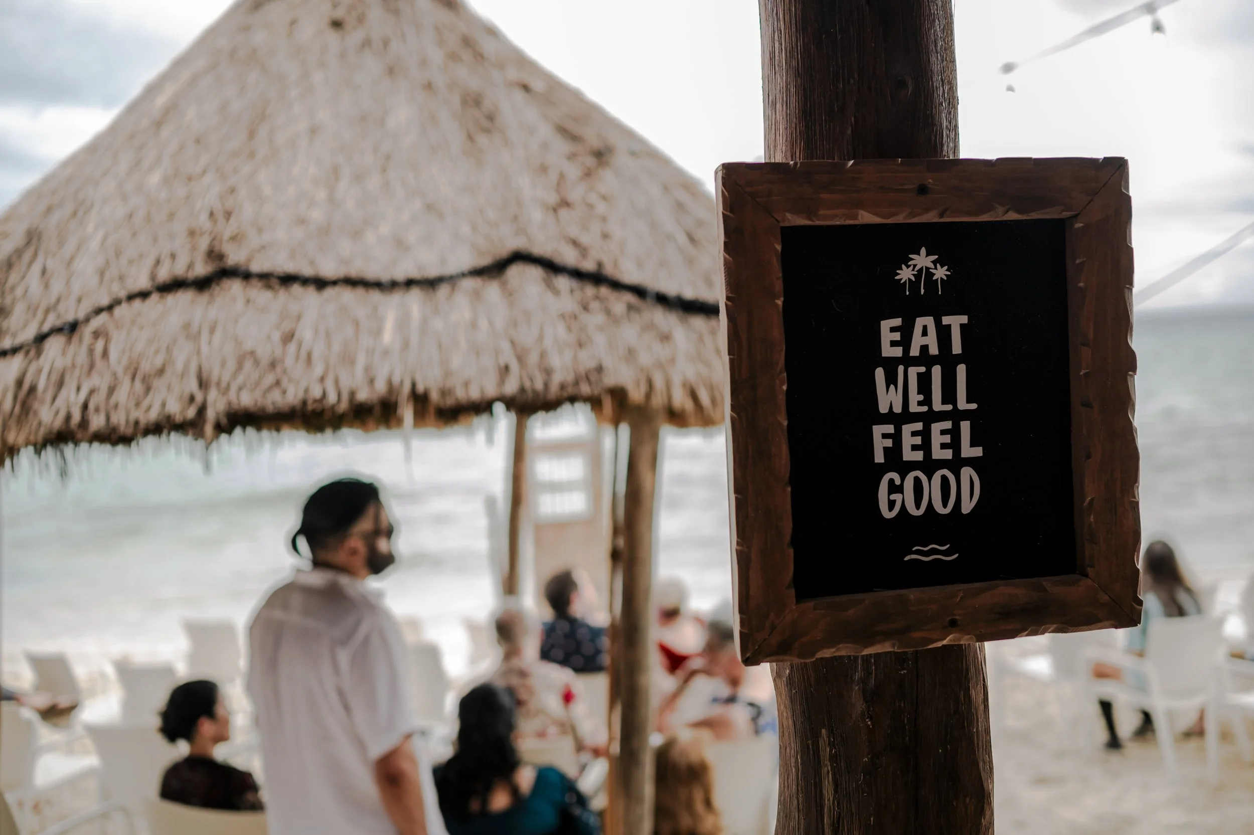 Beach scene with a wooden sign reading 'Eat Well Feel Good' and a thatched umbrella with people sitting at tables by the ocean.
