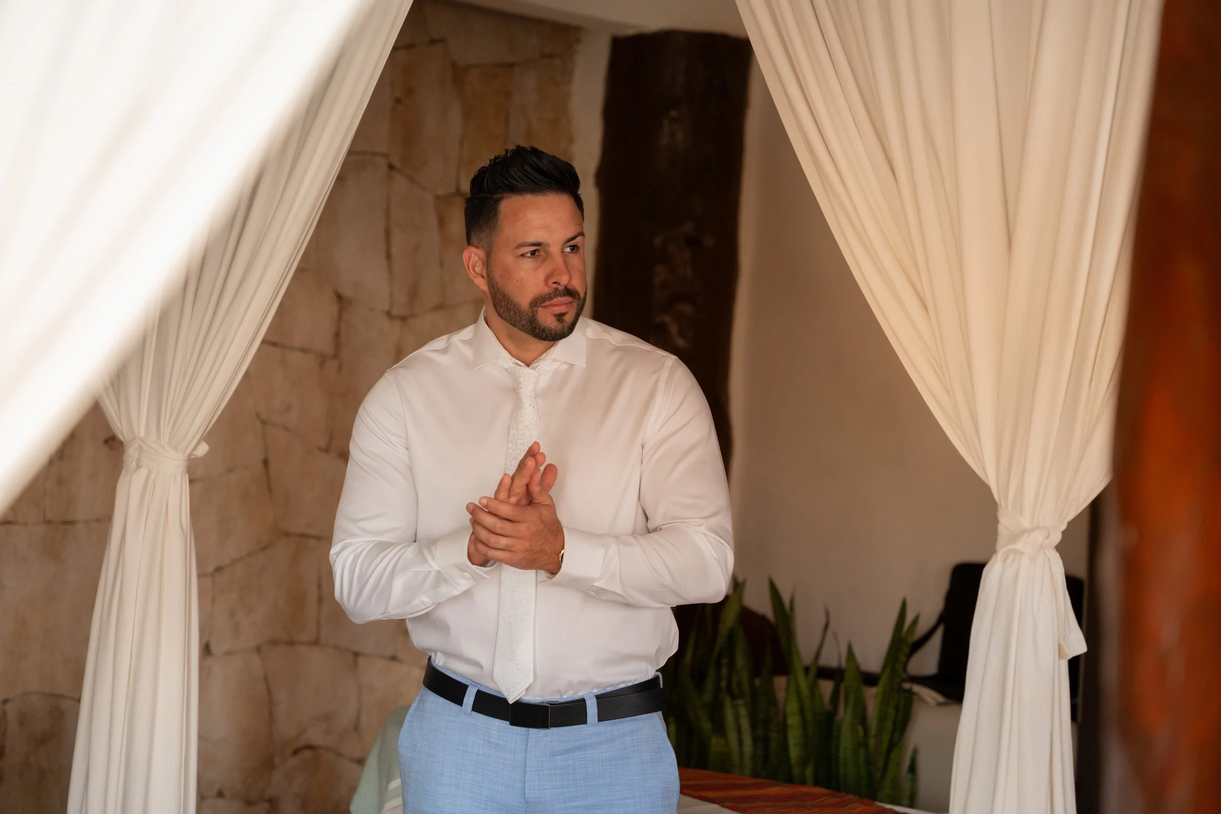 A man with dark hair, beard, and mustache standing inside a gazebo with white curtains, wearing a white dress shirt, light blue pants, and a white tie, looking thoughtfully to the side.