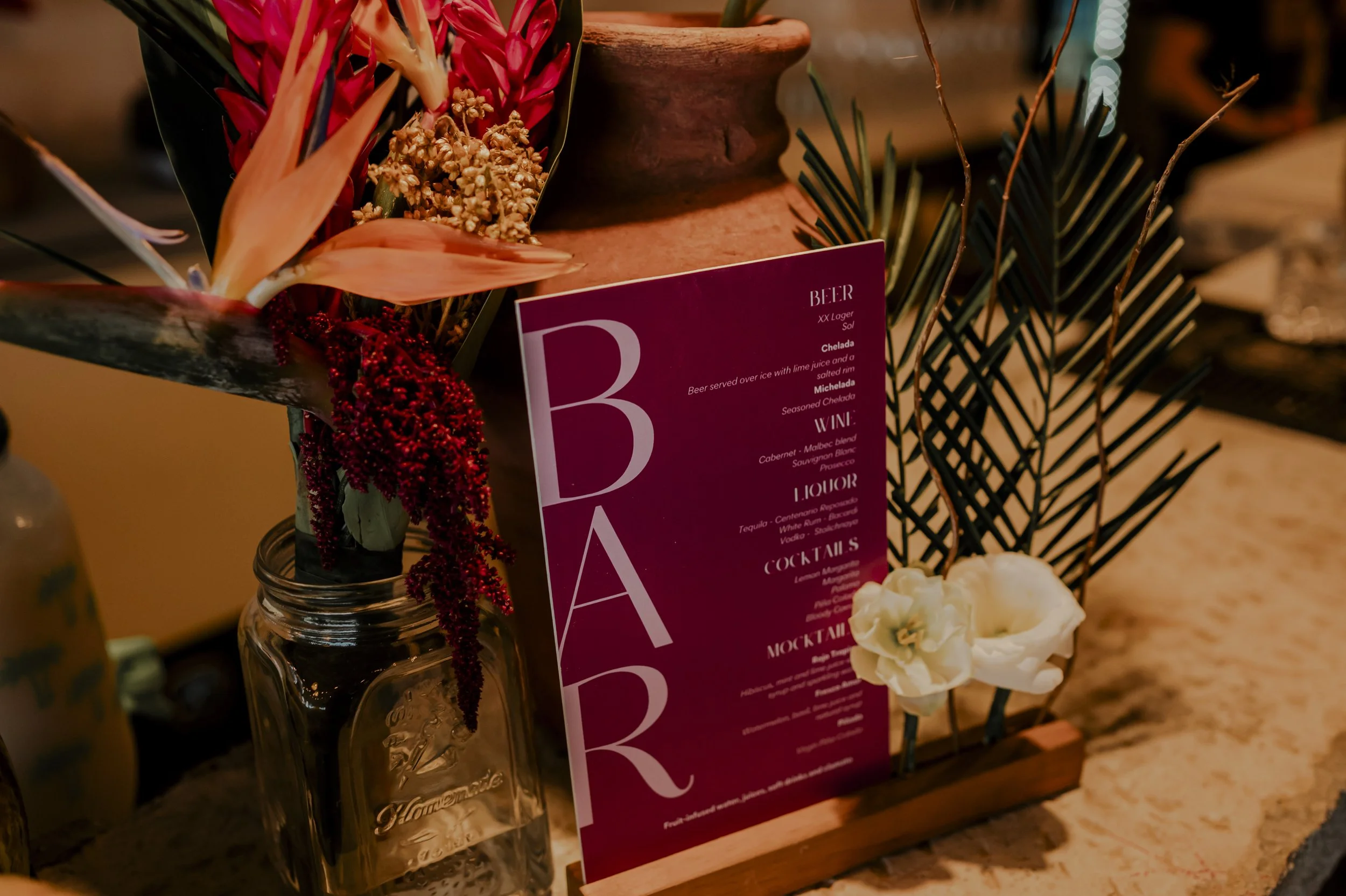 Decorative table arrangement with flowers, a glass jar, and a menu sign for a bar with drinks categories.